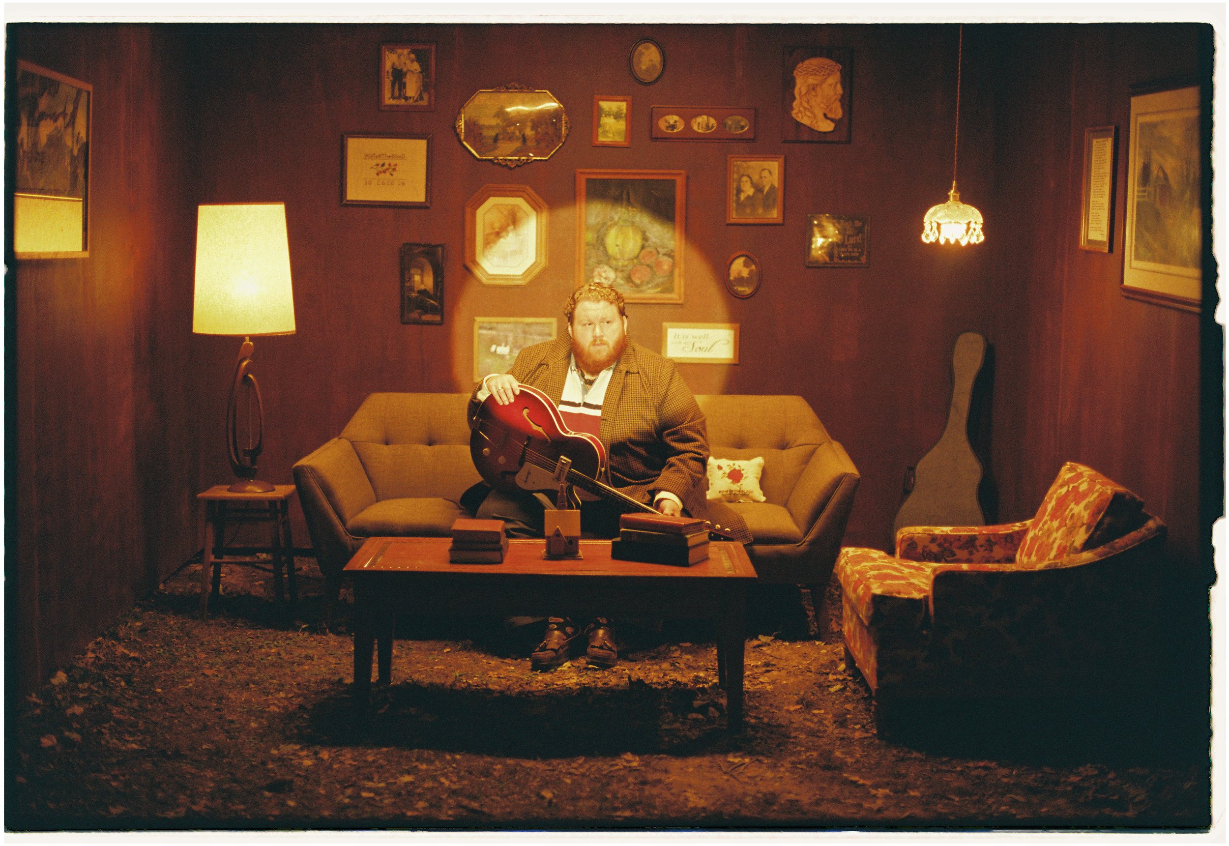 A man with curly hair and a beard sits on a mid-century style brown sofa holding a red electric guitar. The room has wood-paneled walls decorated with various framed pictures and mirrors. There is a standing lamp to the left, a small side table, and a framed pillow on the sofa. To the right, there is a floral armchair and an acoustic guitar case standing in the corner. A hanging lamp provides warm lighting.