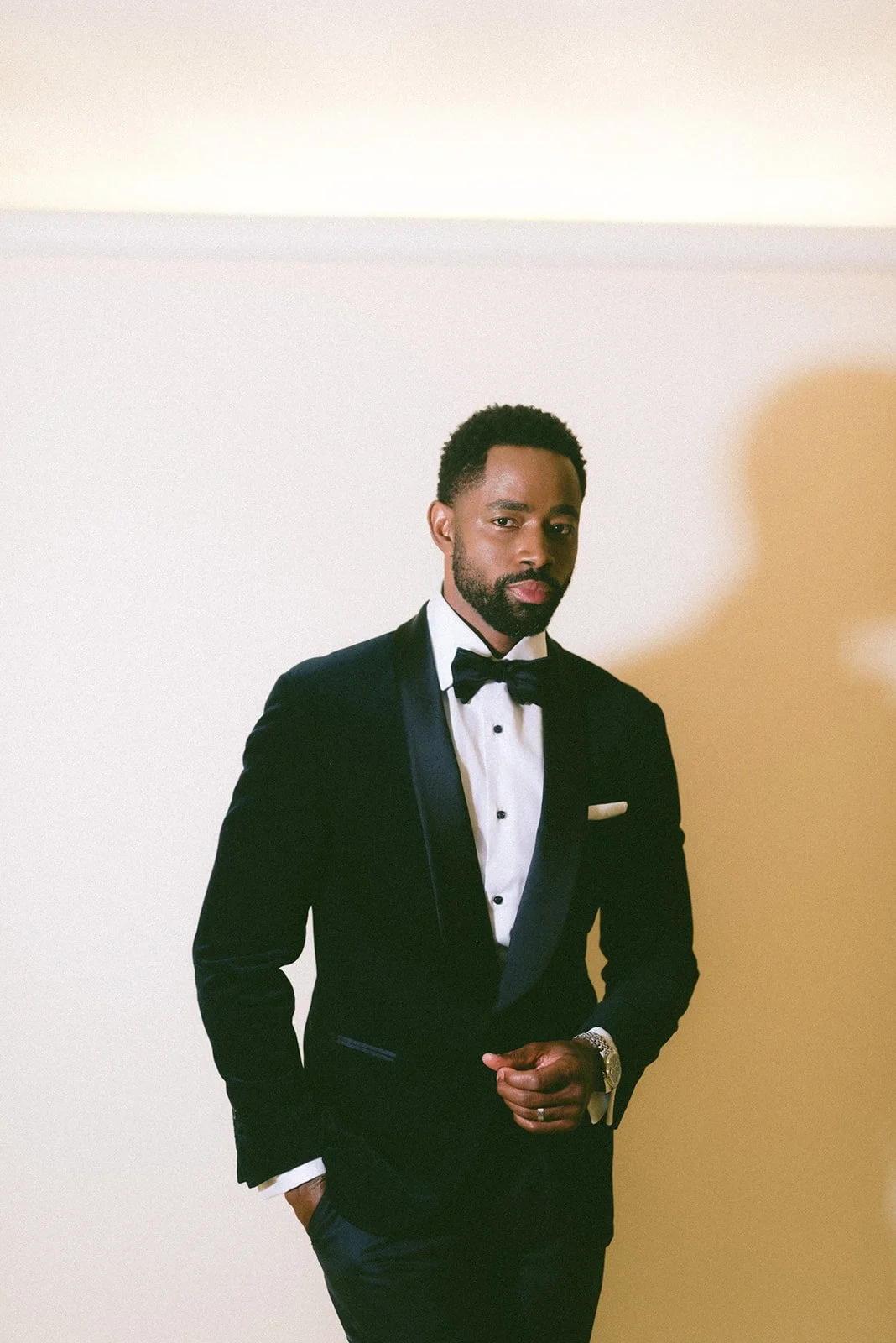 A man dressed in a black tuxedo with a bow tie, standing against a plain beige background.