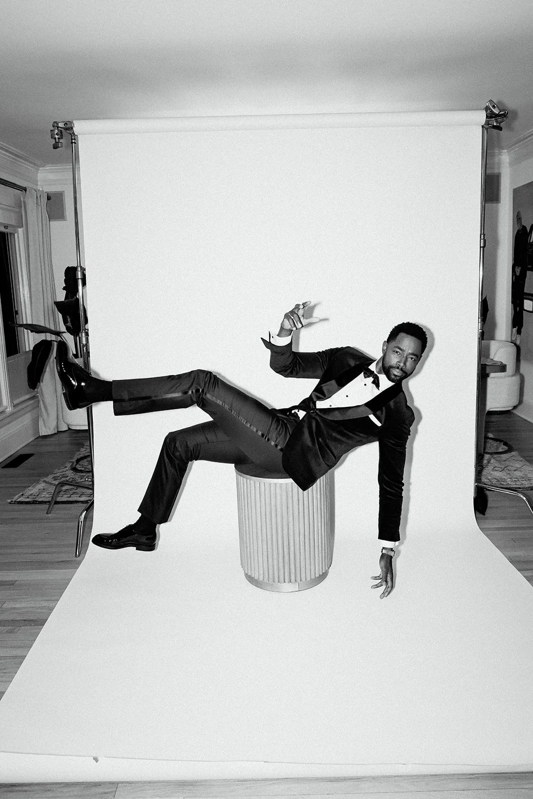 A man in a tuxedo lying on a cylindrical stool on a photography backdrop, with his body extended and one hand touching the floor, posing for a photoshoot in a home setting.