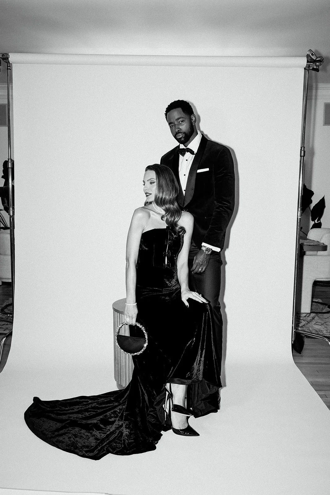 Black and white photo of a man in a tuxedo standing behind a seated woman in a strapless gown, both looking stylishly posed in a photo studio.