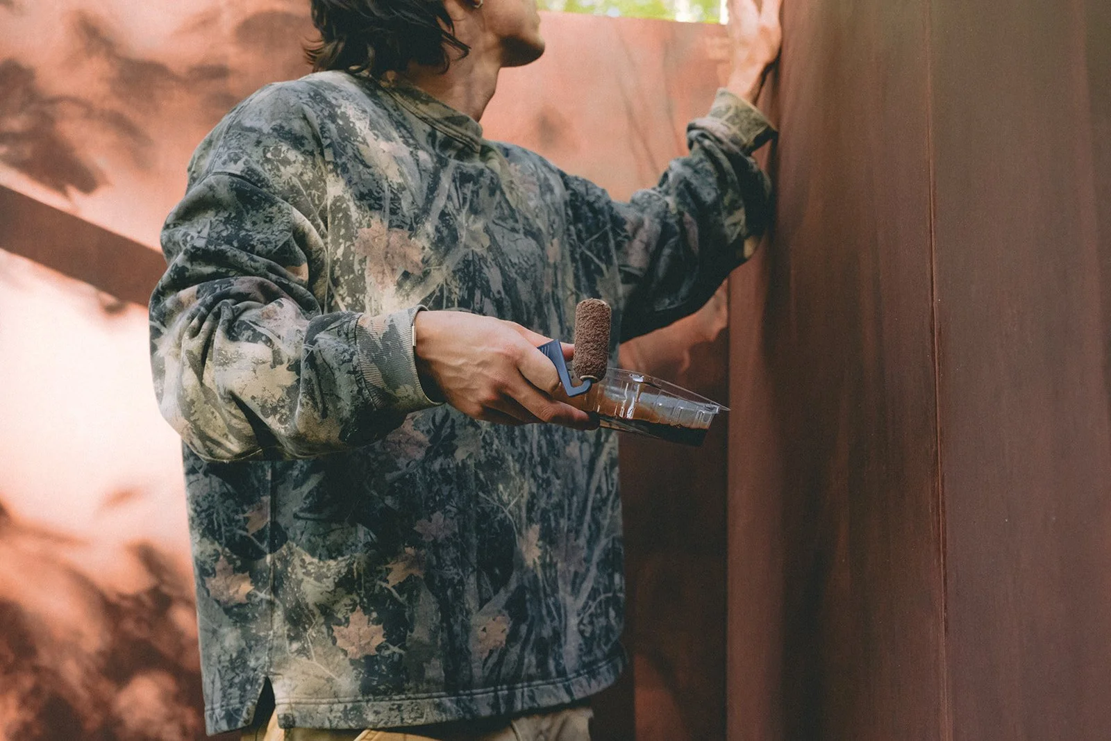 Person painting a wall with a roller paintbrush, wearing a camouflage jacket.