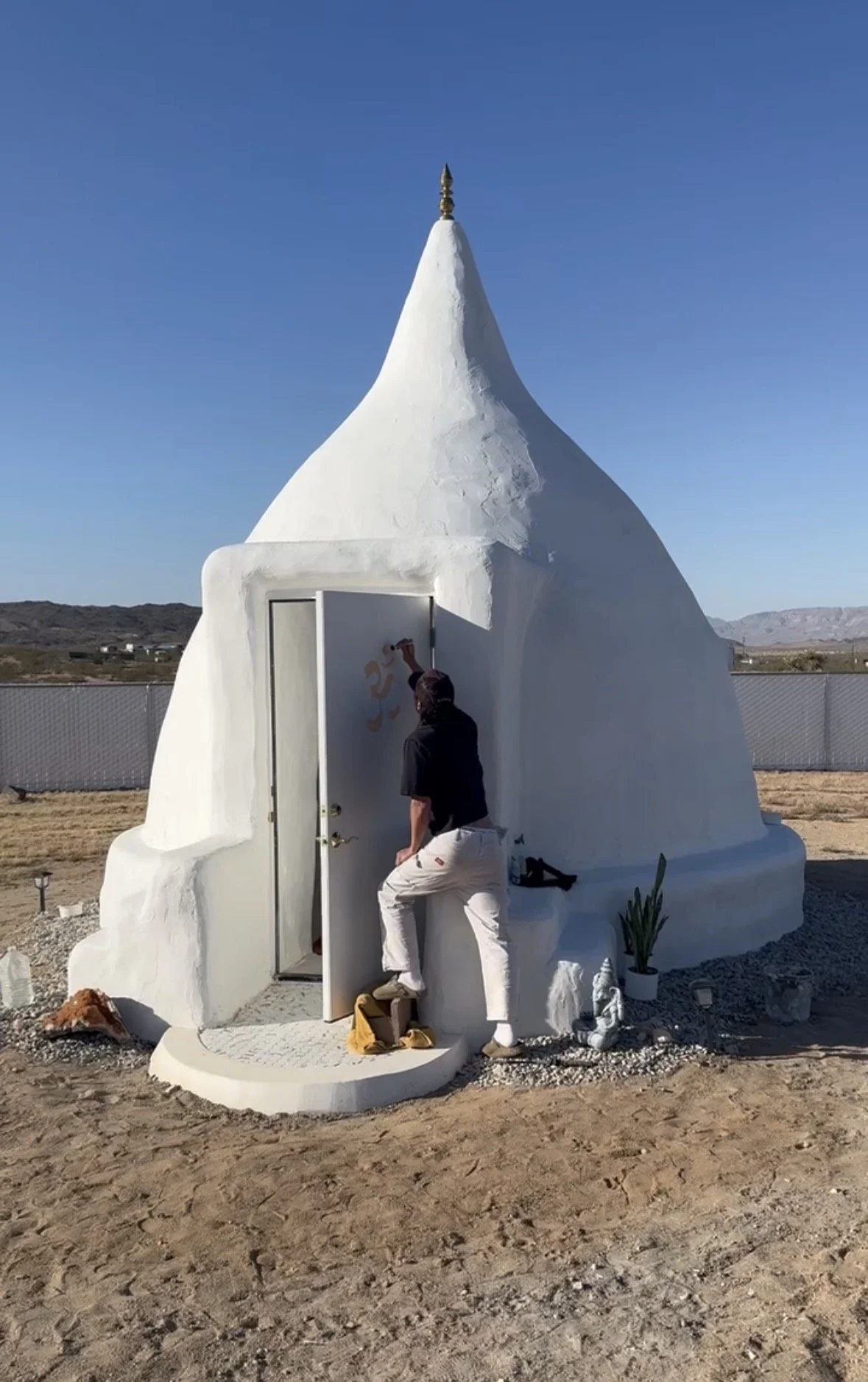 A person painting the Om symbol on a small, white, domed chapel-style building, resembling a miniature church or shrine, outdoors with a clear blue sky.