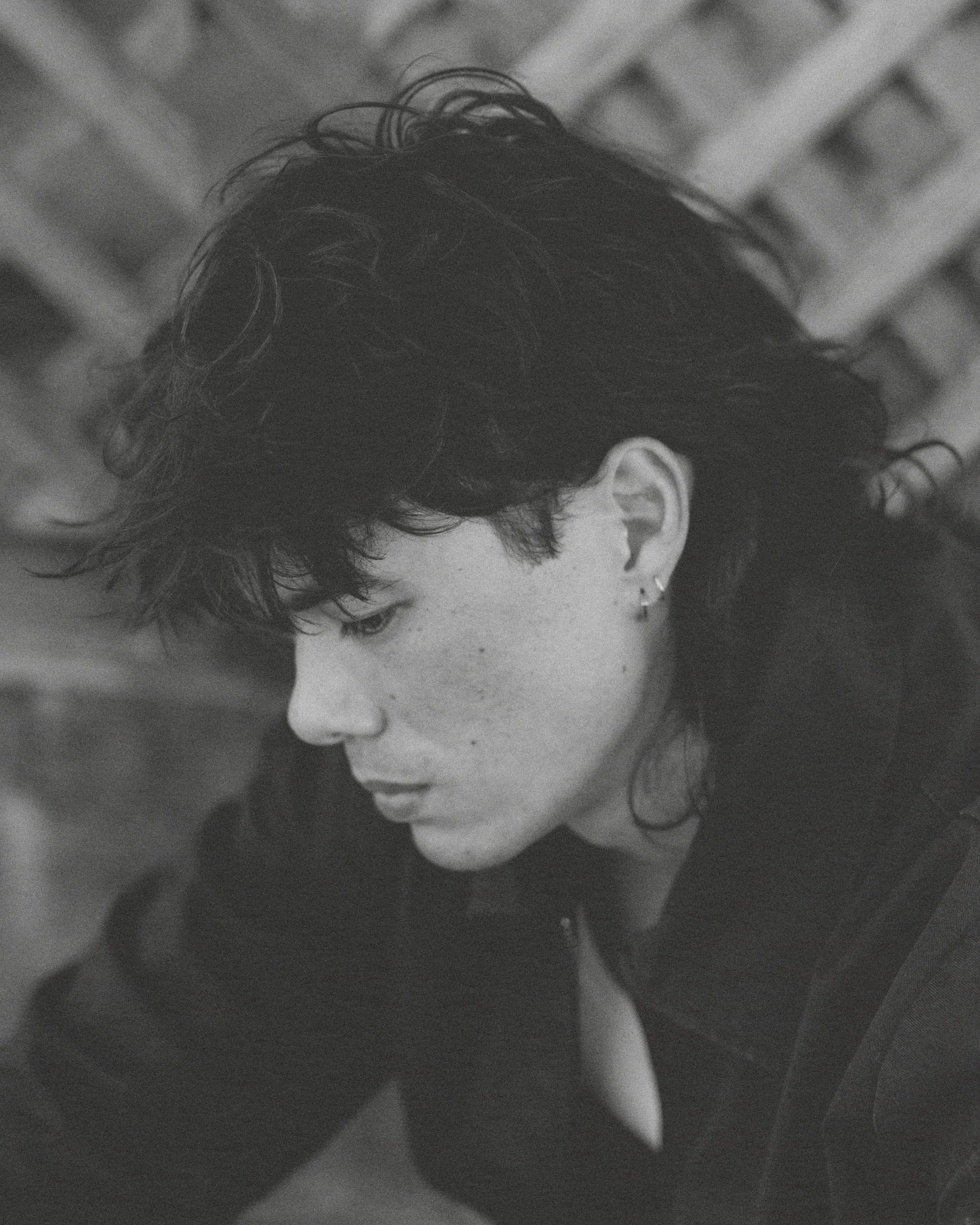 Black and white photo of a woman with short wavy hair, earrings, and freckles, looking downward with side profile visible, against a blurred background.