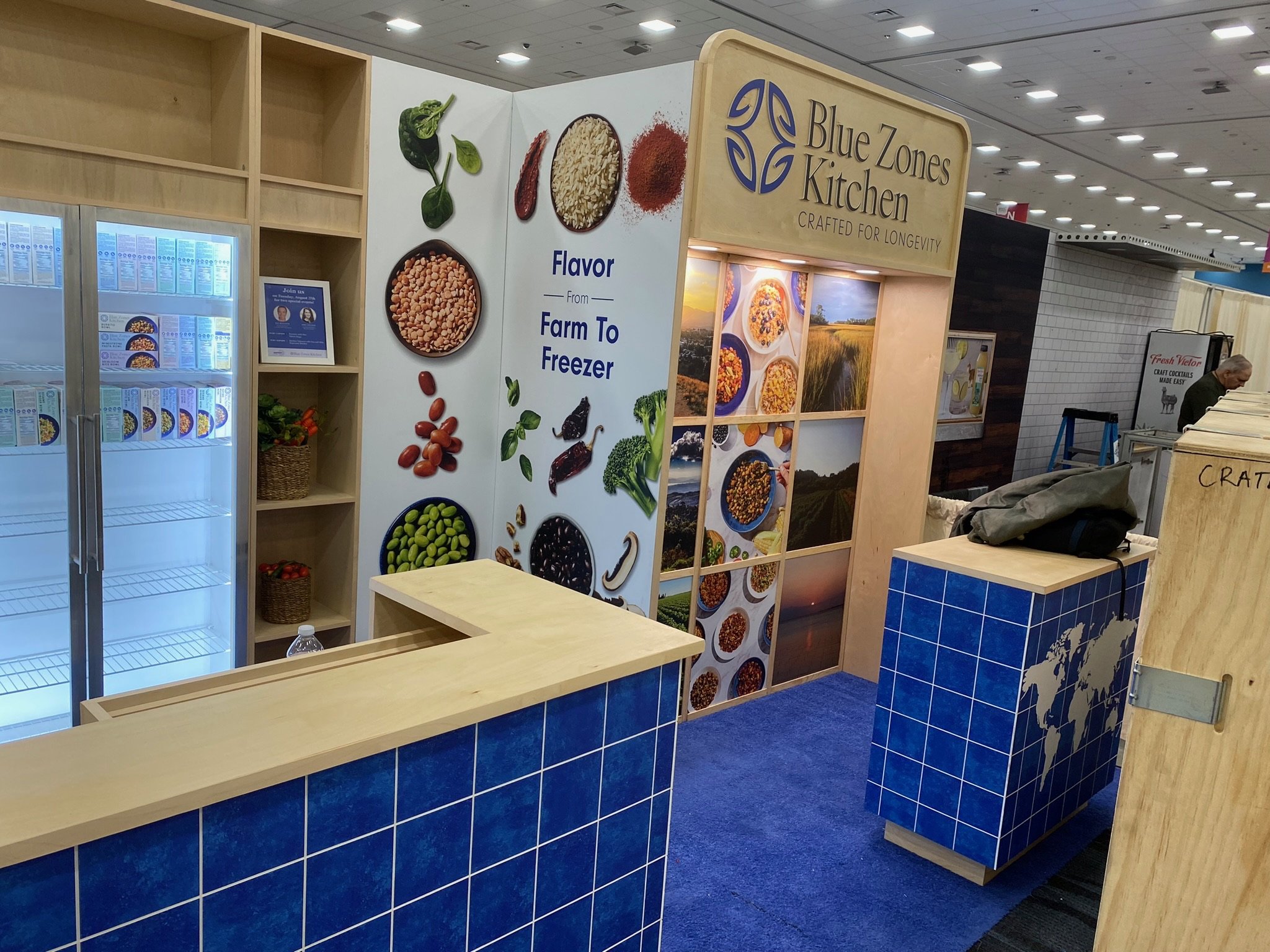 Booth for Blue Zones Kitchen at an exhibition, featuring a display wall with images of various foods and scenic landscapes, a blue tiled counter, and a refrigerator stocked with products.