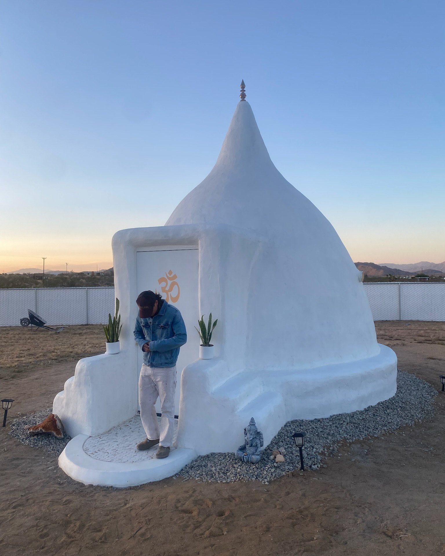 A white, dome-shaped building resembling a small temple or shrine with a tall, pointed spire on top, two potted cacti on steps at entrance, a person in a denim jacket and white pants standing in front, and a small statue of Ganesha nearby, set against a sunset sky.