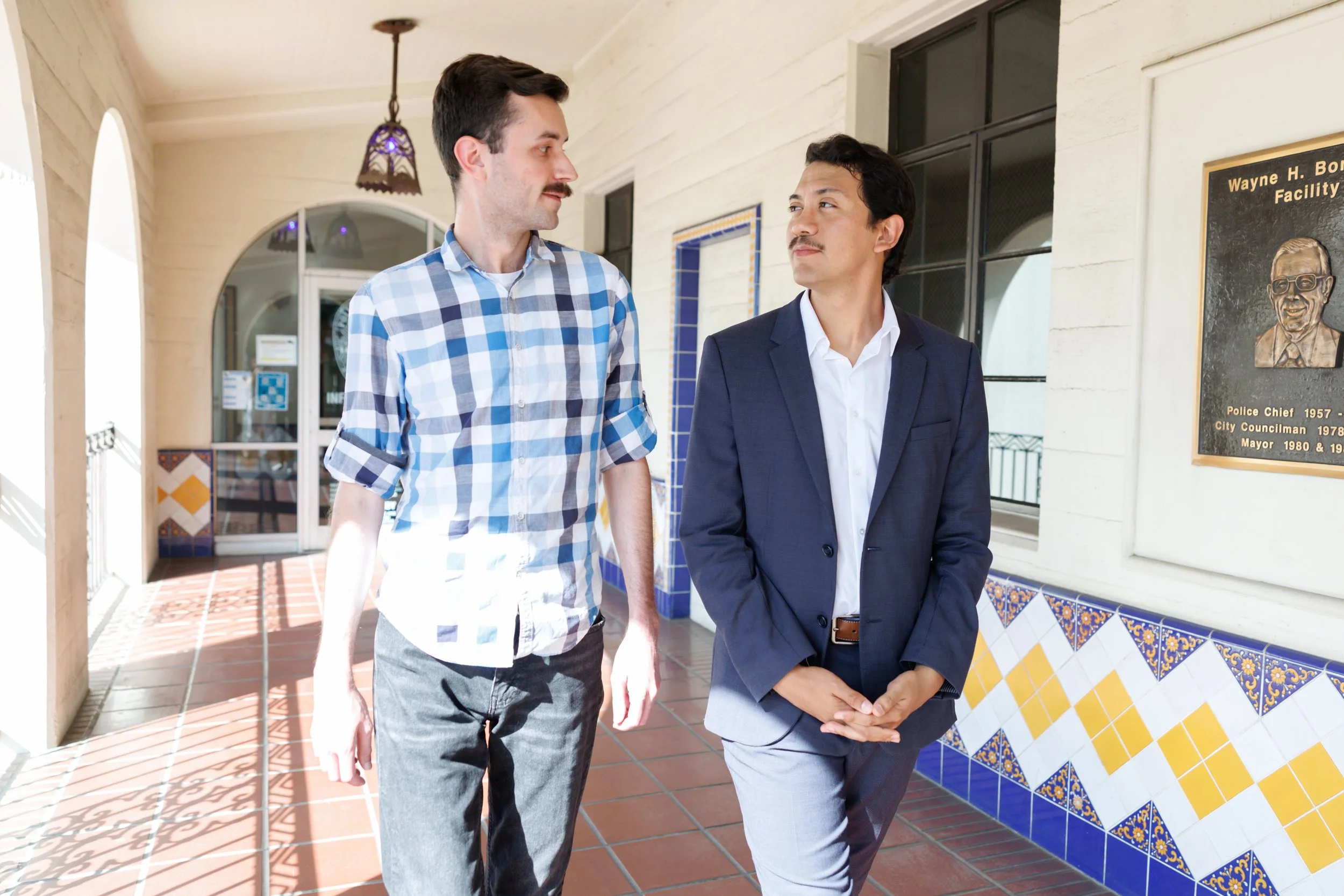 Two men in business attire walking past a wall with a plaque and decorative tiles, engaging in conversation outside a building.