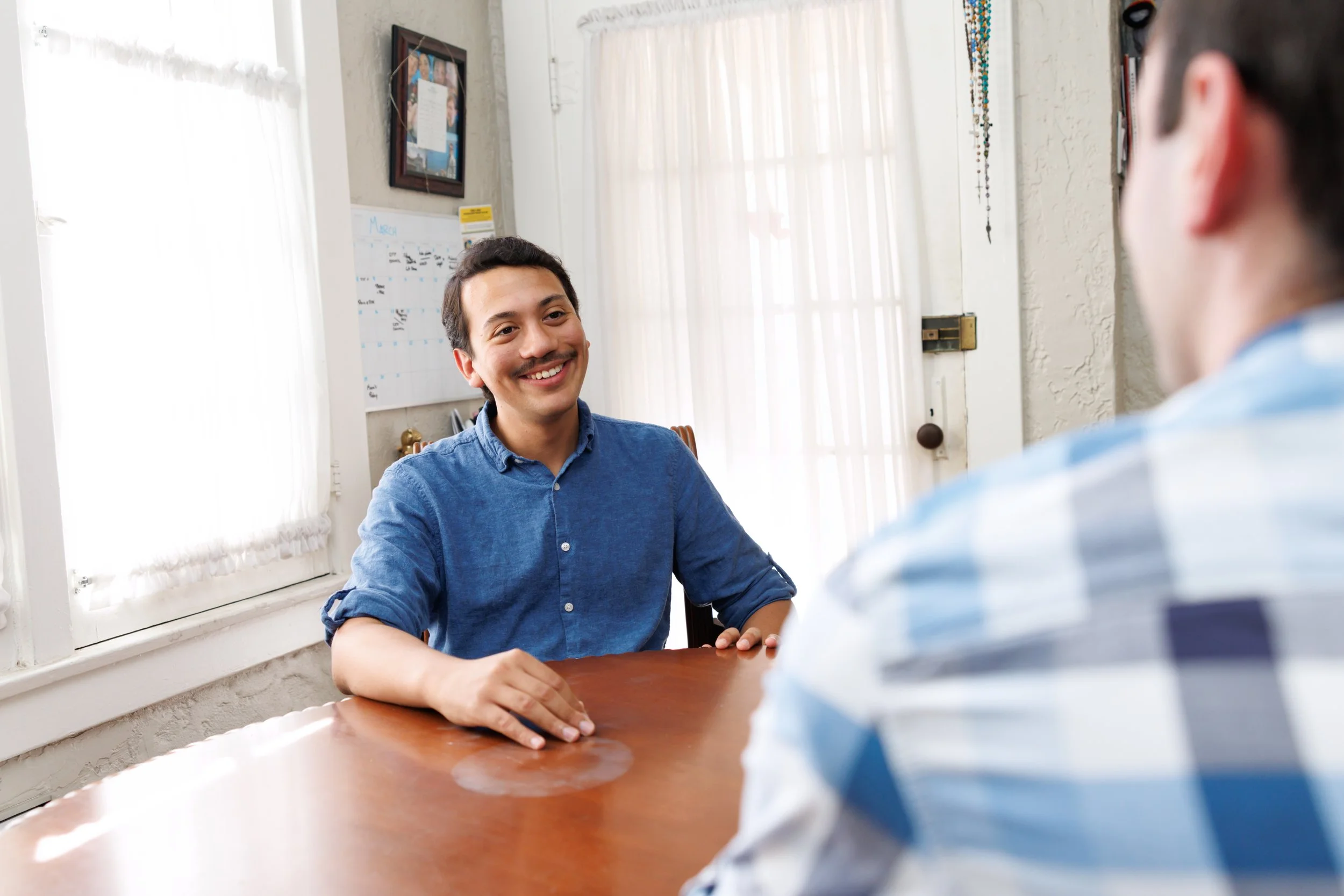 Two men sitting at a wooden table, one smiling and wearing a blue shirt, the other partially visible and facing away, in a bright room with white curtains and framed photos on the wall.