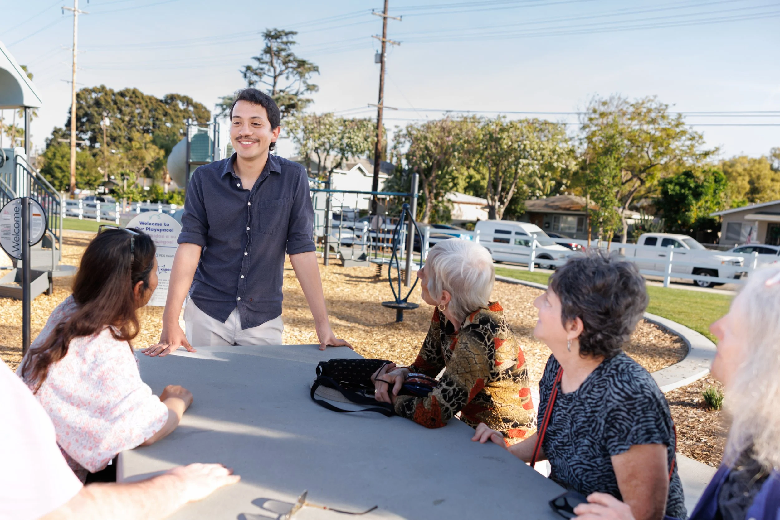 A man with dark hair and a mustache is standing and smiling at a table among a group of elderly women outdoors in a park or playground area. The women are seated and listening attentively. There are trees and parked cars in the background.