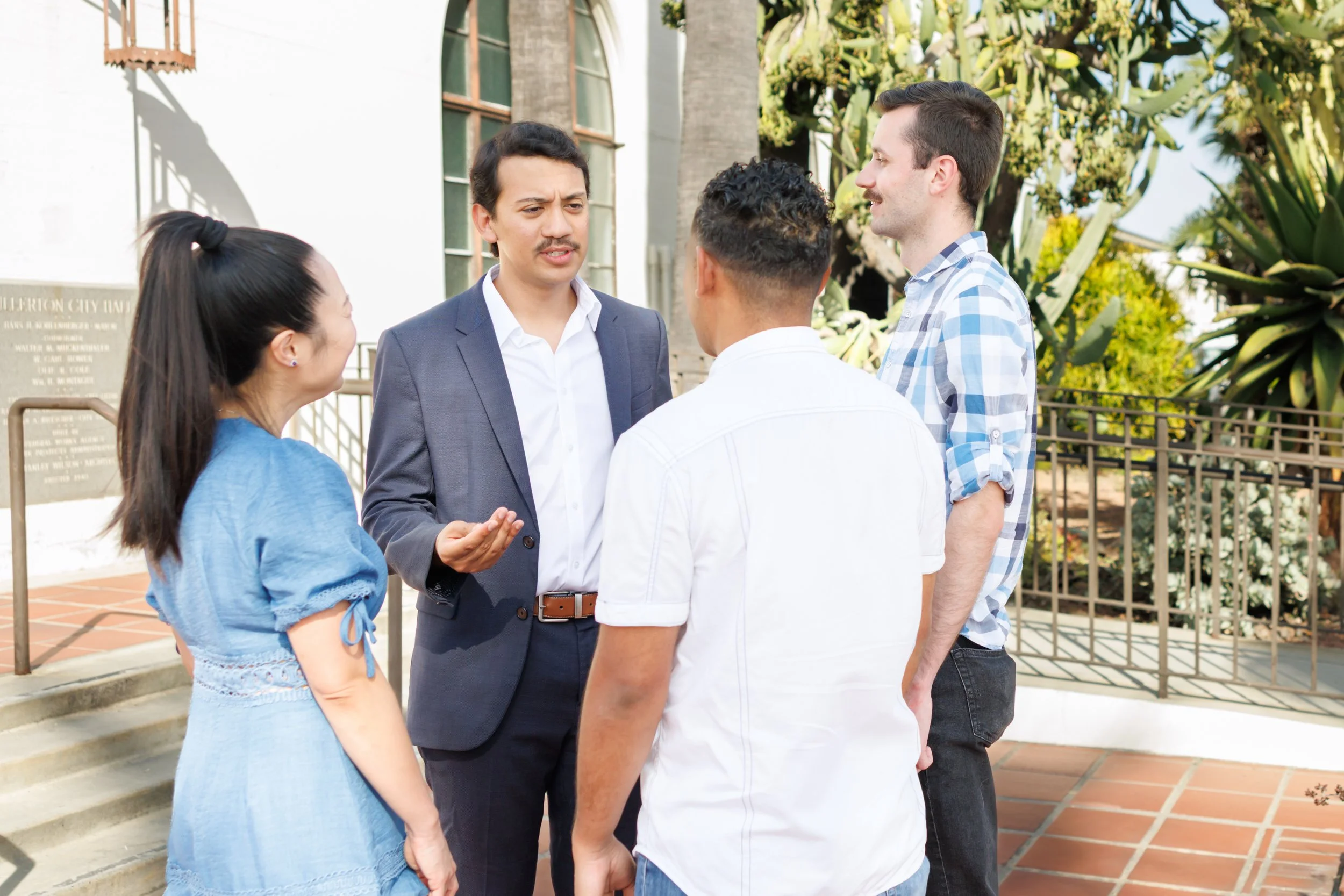 Five people engaged in a conversation outside a building, with greenery and a tree in the background.