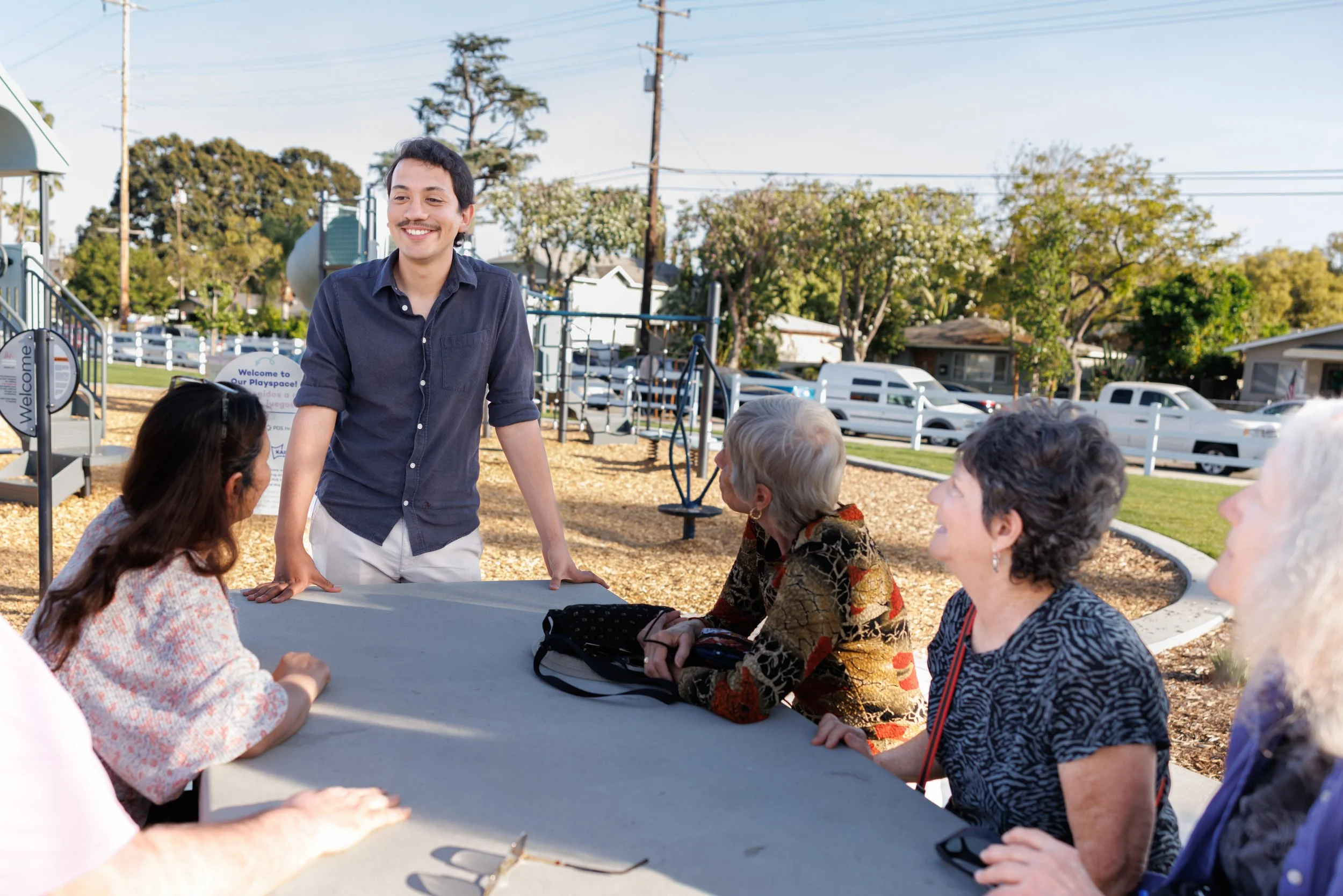 A young man with dark hair and a mustache stands smiling at an outdoor gathering of older women seated around a table, with trees, parked cars, and a playground in the background.