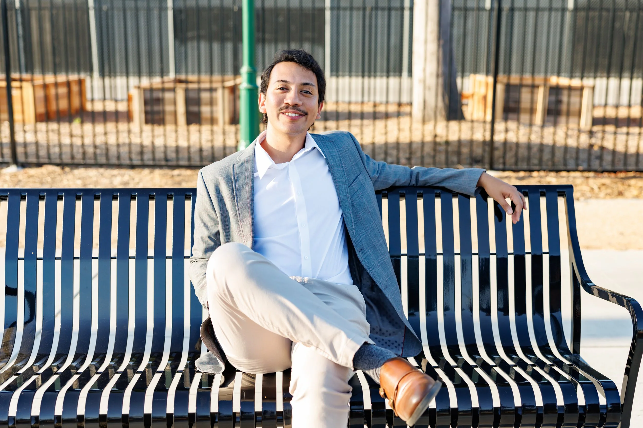 A young man with dark hair and a mustache sitting on a black metal park bench outdoors, smiling, wearing a light grey blazer, white shirt, beige pants, and brown boots, with a background of a fenced area and wooden benches.