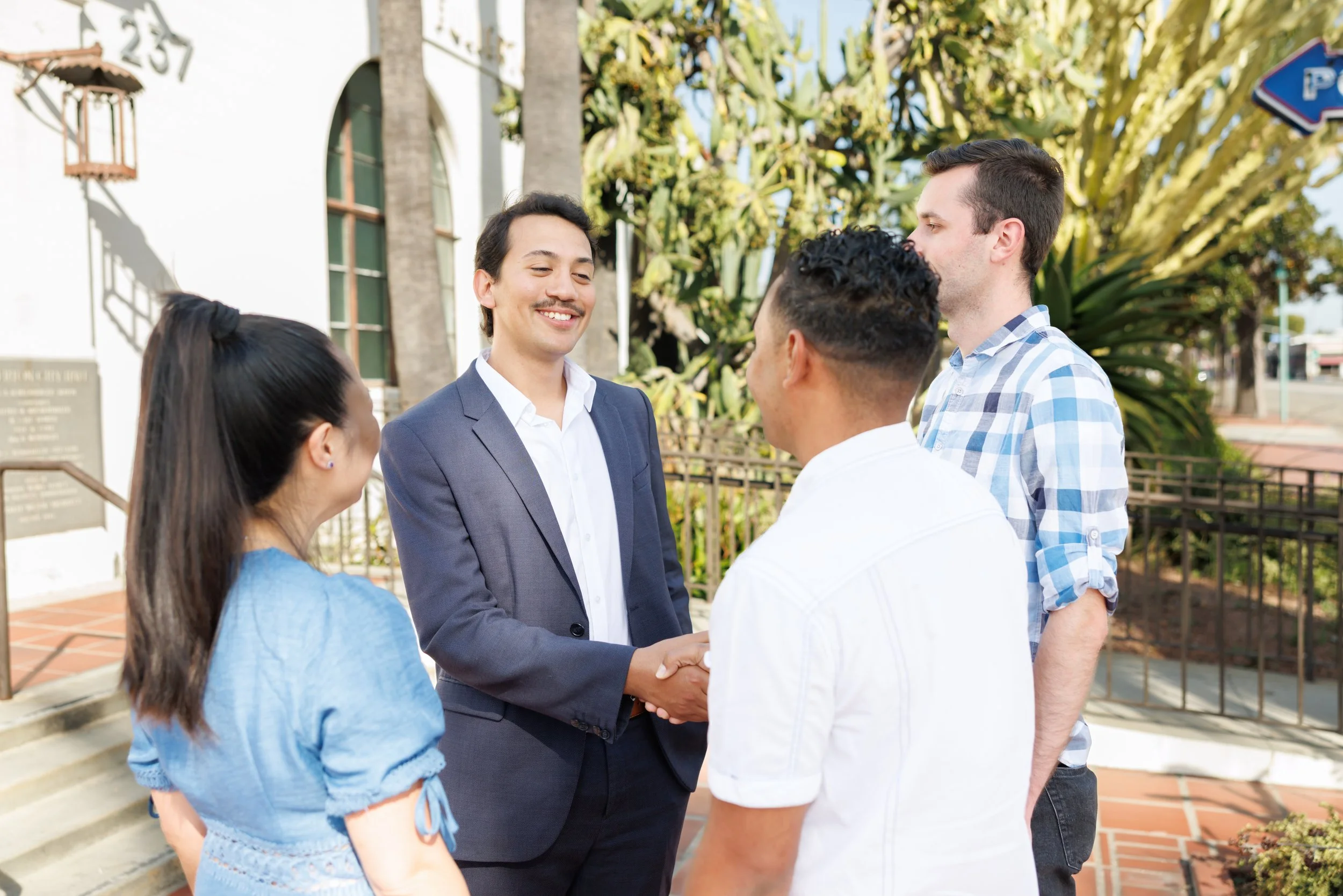 A group of five people, including a man in a suit and women, are outdoors in front of a building, engaged in a conversation. The man in the suit is shaking hands with one of the women.