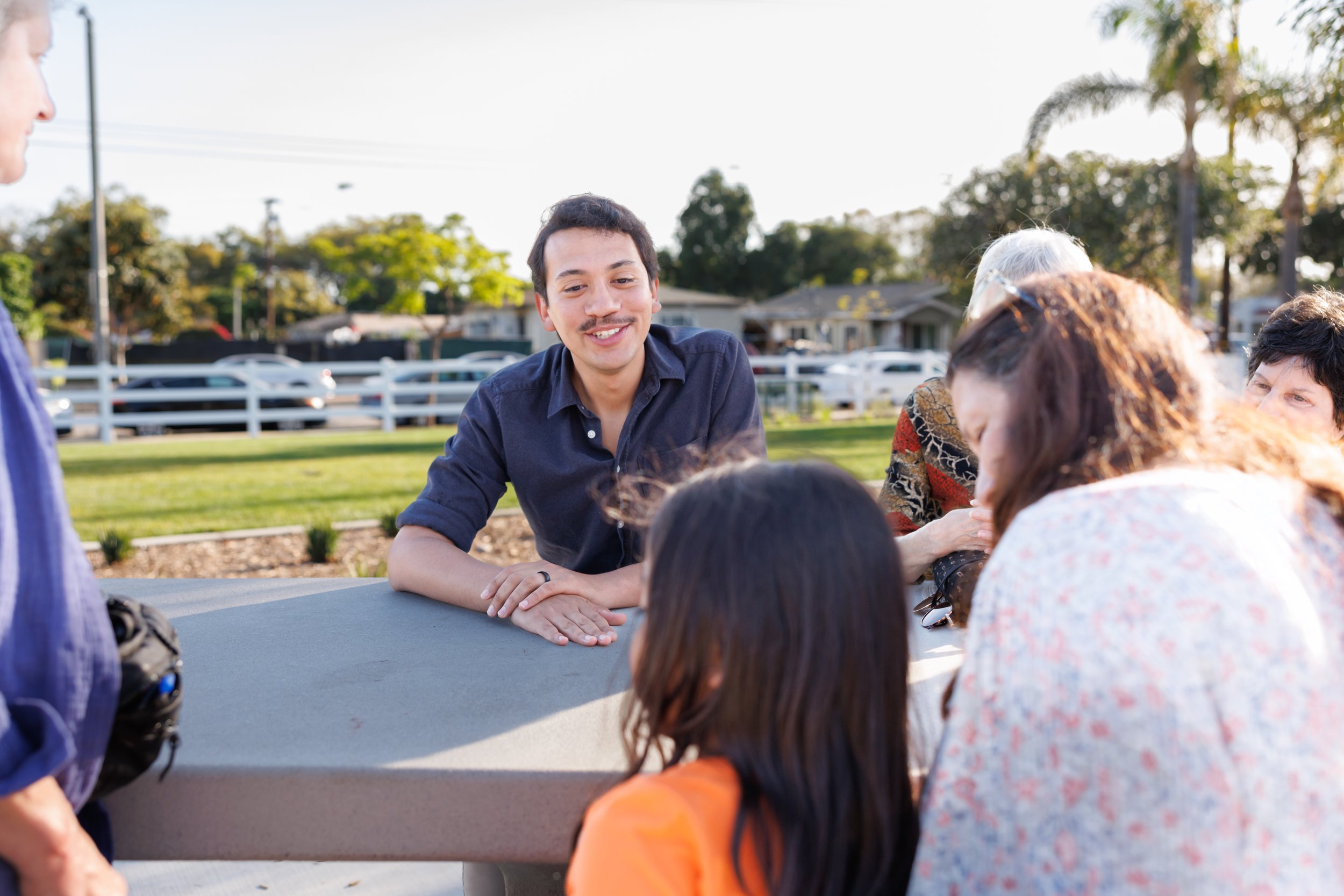 A young man with dark hair and mustache smiling and leaning on a table, engaging with a group of diverse people outdoors in a park, with trees, houses, and cars in the background on a sunny day.