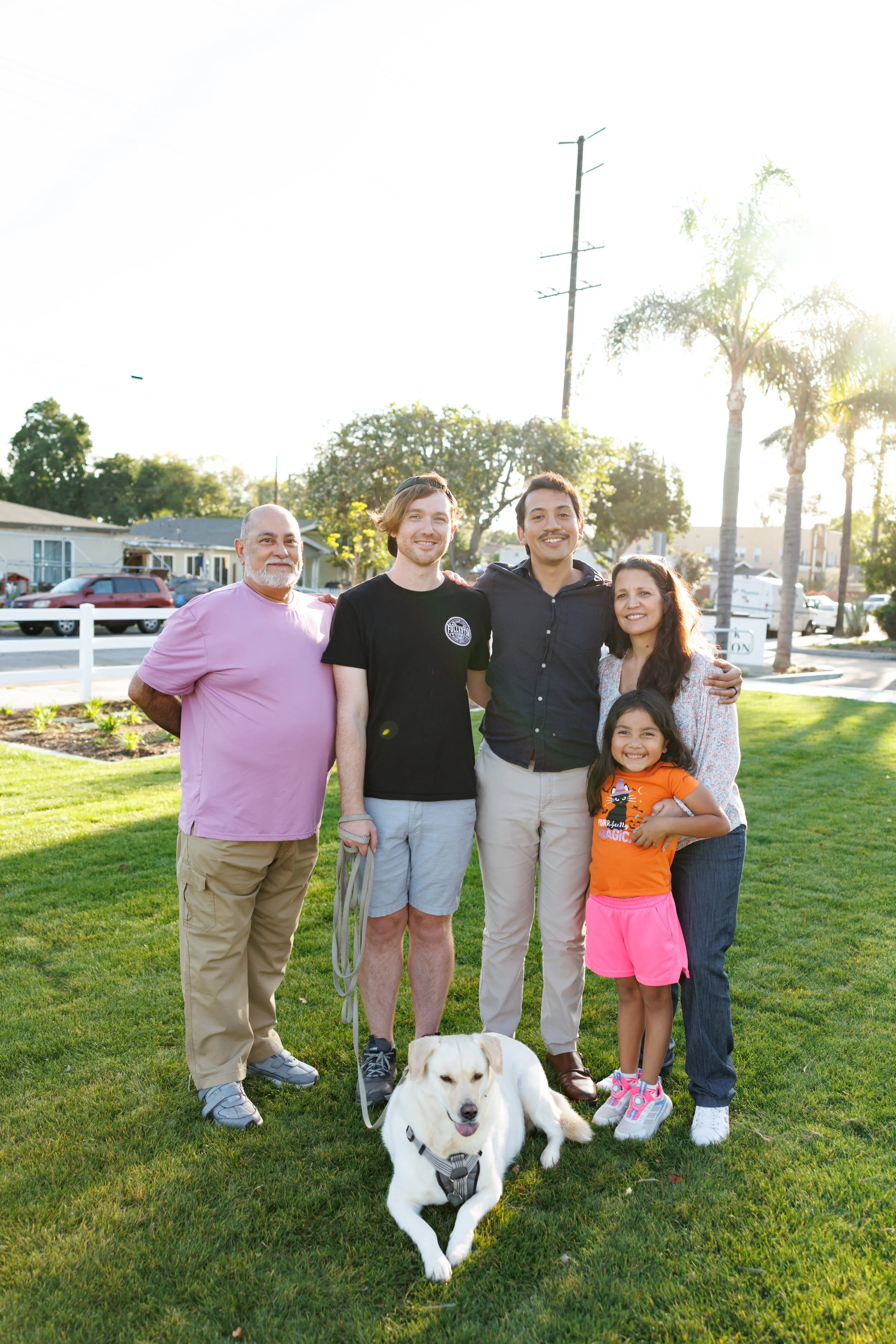 A diverse family group outdoors in a park-like setting with four adults, two children, and a white dog. The sun shines brightly behind trees and palm trees in the background.