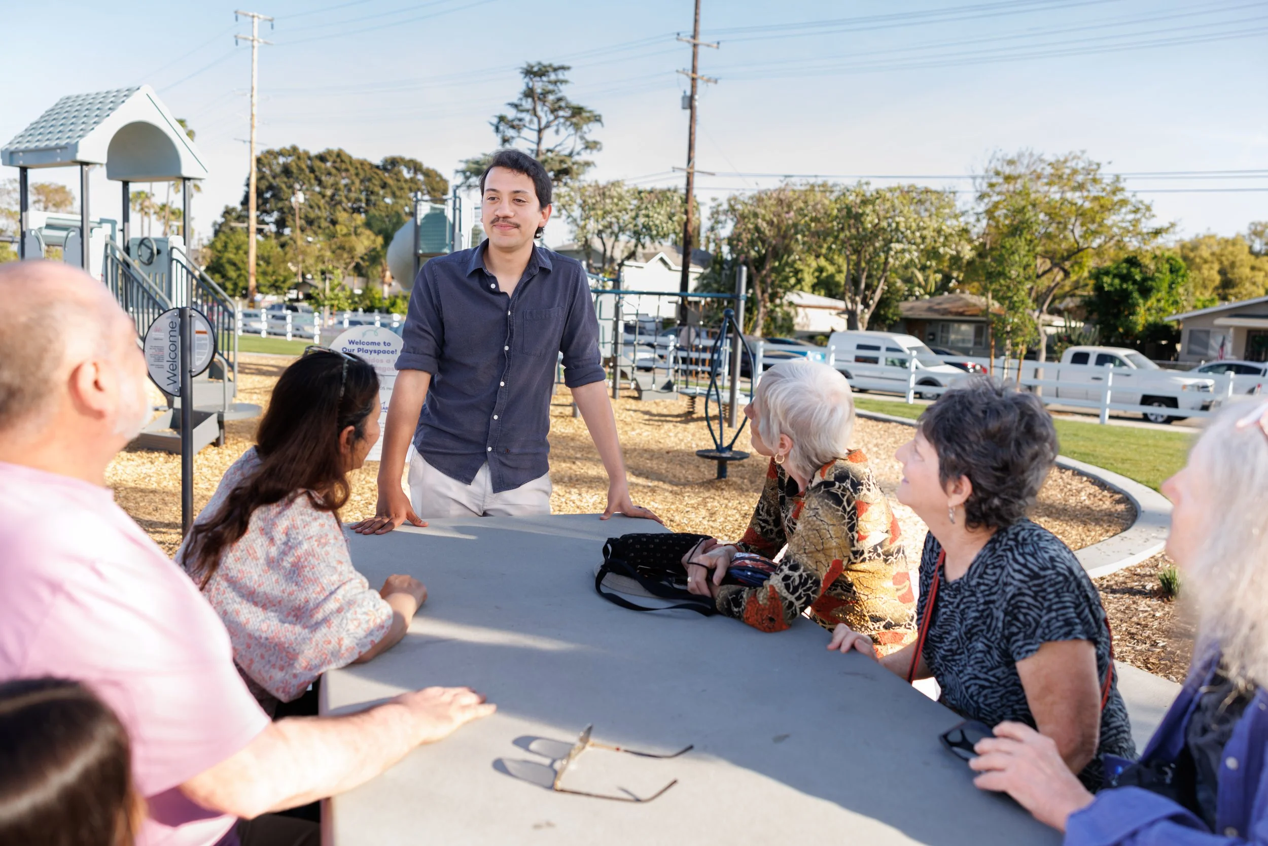 A young man with dark hair and a mustache stands at a table talking to a group of elderly people seated around it at a playground. The playground has a slide and blue azalea hedge in the background with parked cars and houses beyond. The sky is blue 