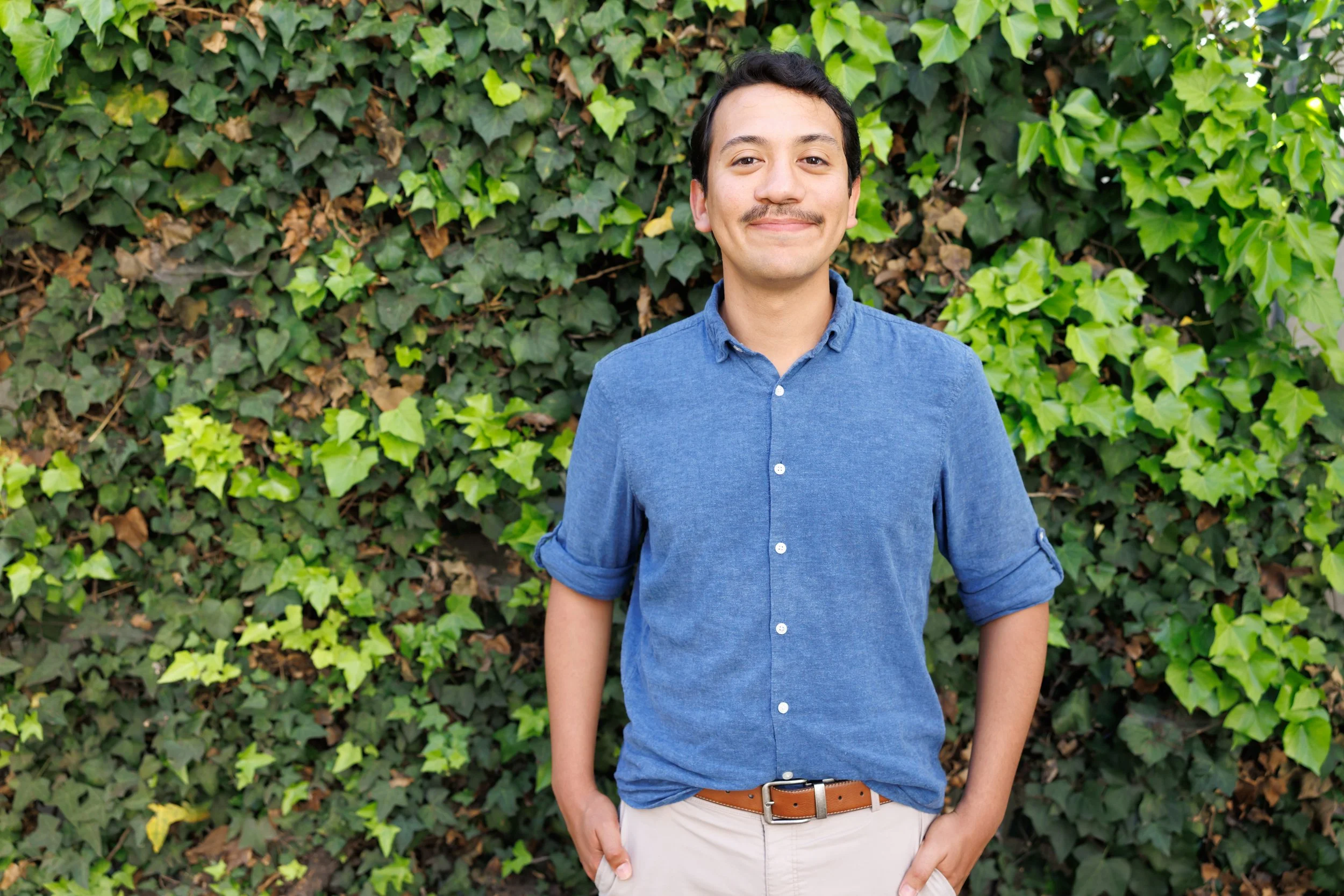 A young man with dark hair and a mustache, wearing a blue button-up shirt and khaki pants, standing in front of a lush green ivy-covered wall, smiling softly with his right hand in his pocket.