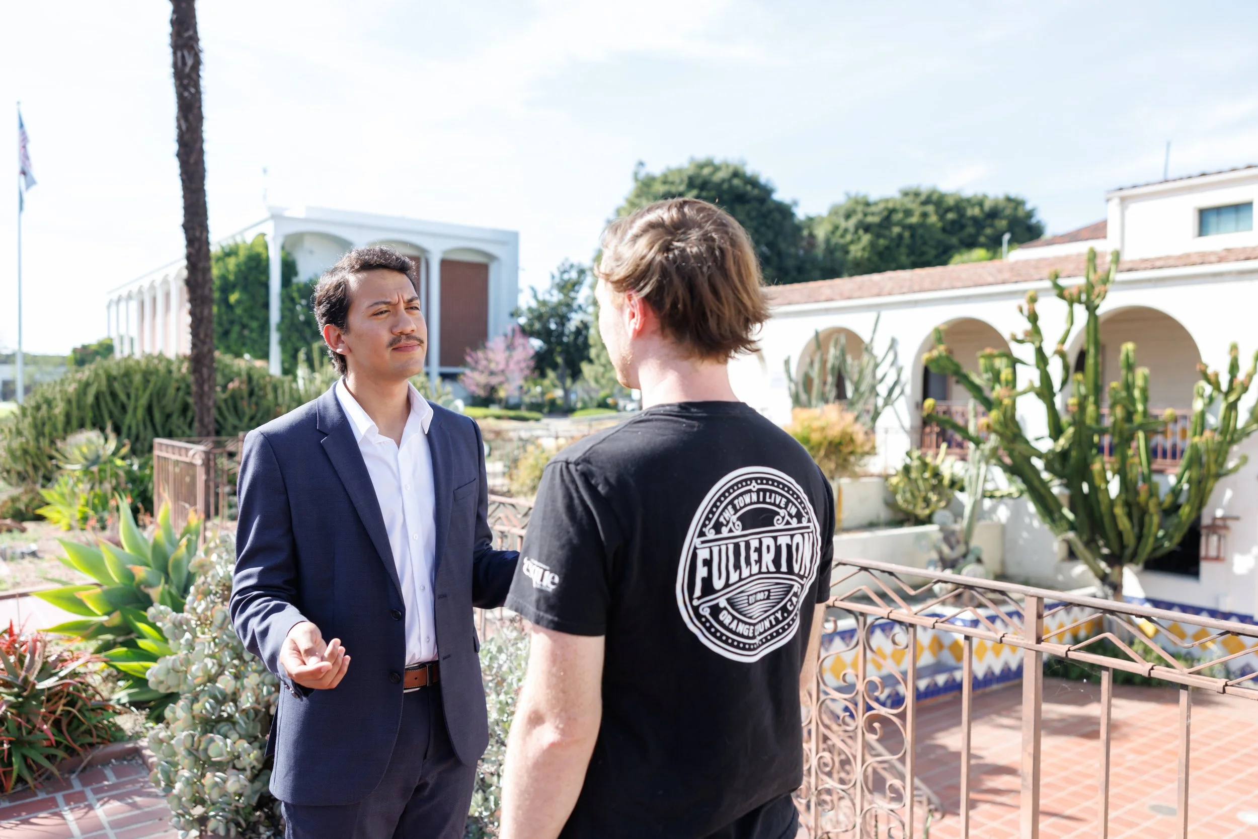Two men interacting outdoors in a garden area with plants and succulents. One man is wearing a navy blue suit, white shirt, and brown belt, and the other man is wearing a black T-shirt with a logo on the back. They appear to be engaged in conversatio