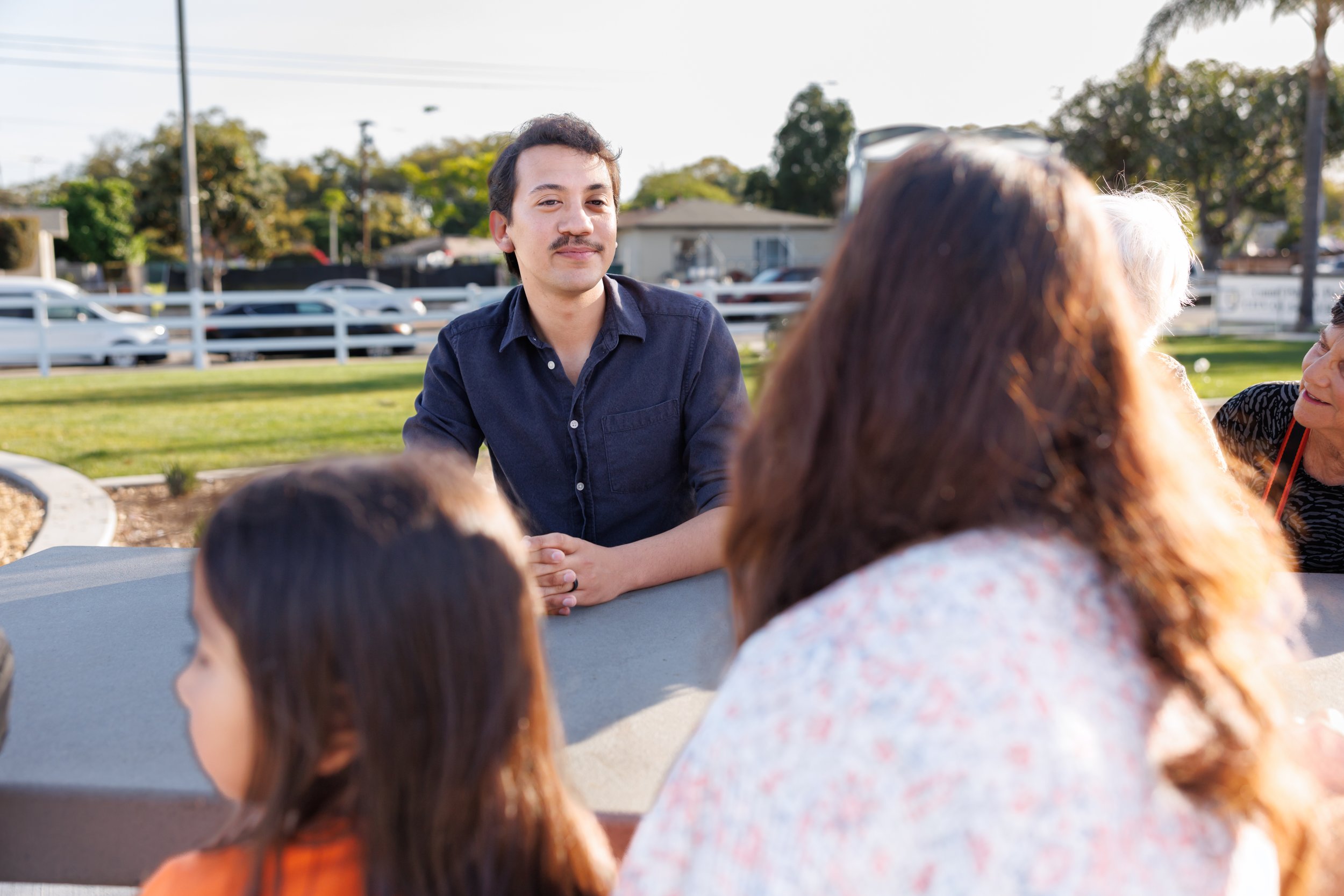 A young man with dark hair and a mustache wearing a navy blue shirt sitting outside at a table, engaging in conversation with a group of people whose backs are to the camera. Background shows cars, trees, and a residential neighborhood.