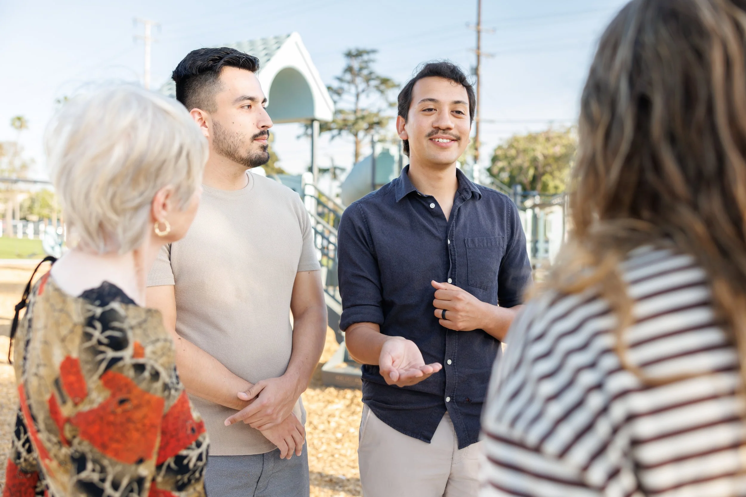A group of people is having a conversation outdoors at a park, with trees and playground equipment in the background.