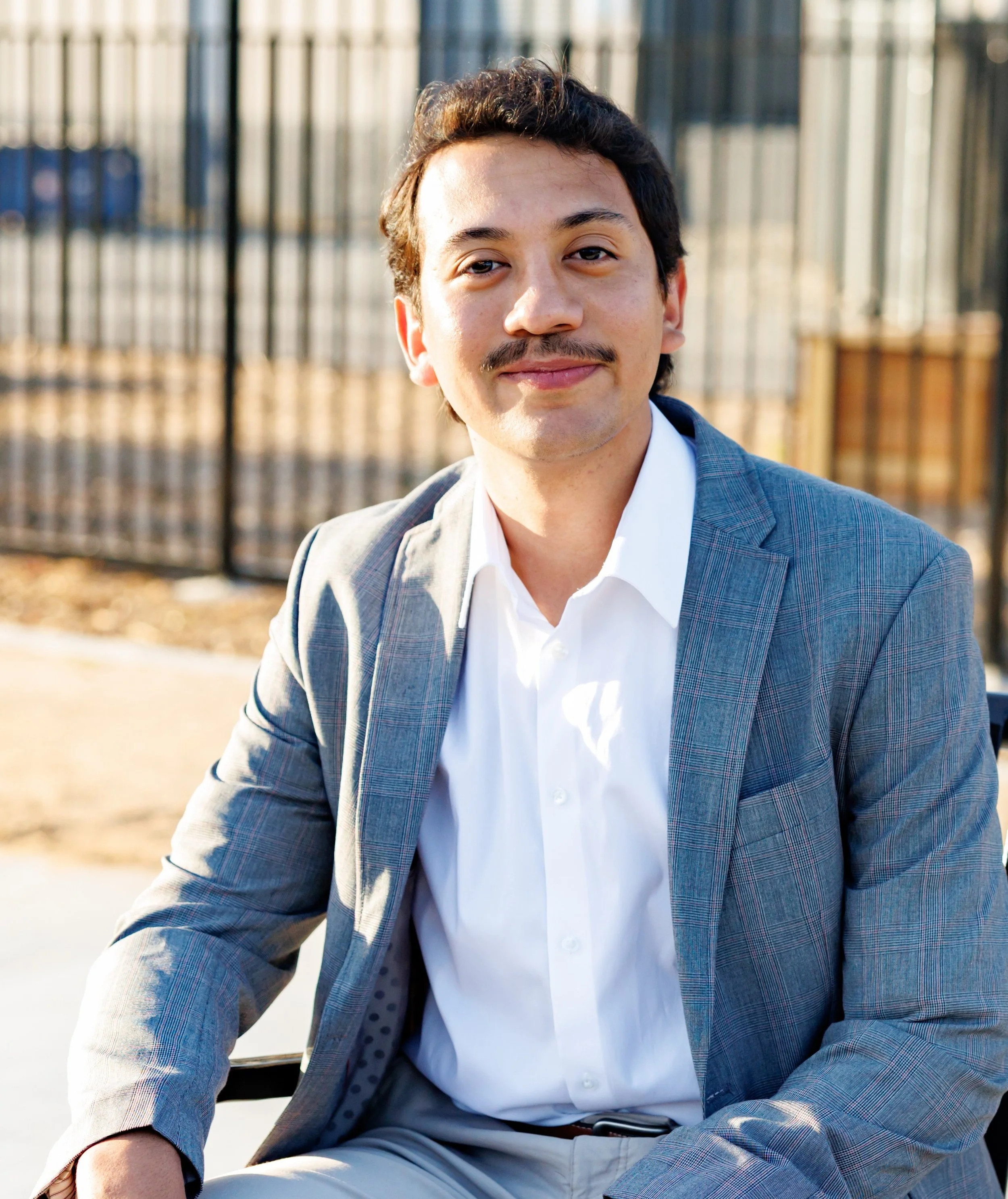 A young man sitting outdoors, wearing a grey plaid blazer, white shirt, and beige pants, with a fence in the background.