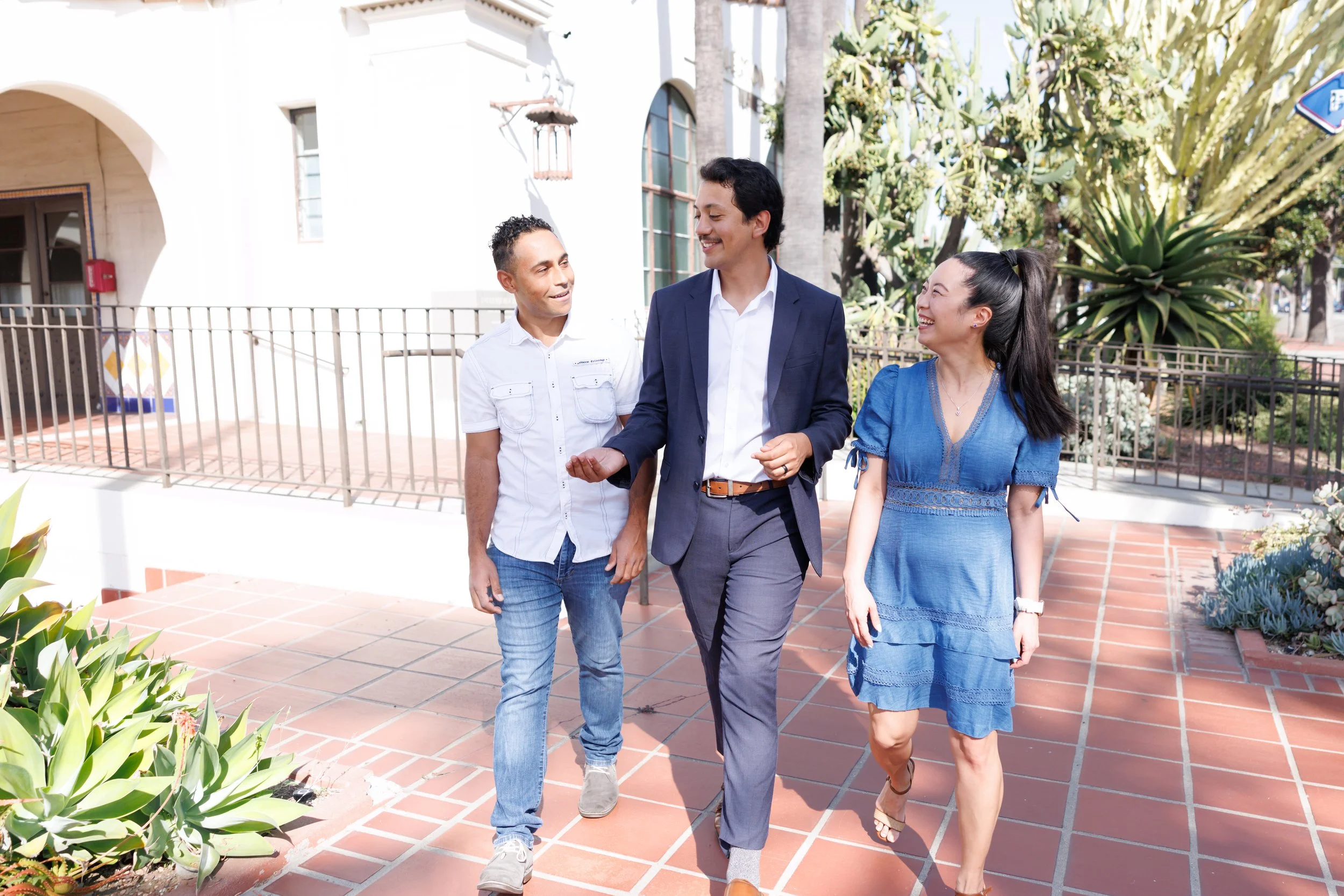 Three people walking together on a sunny sidewalk in an urban area, smiling and engaged in conversation.