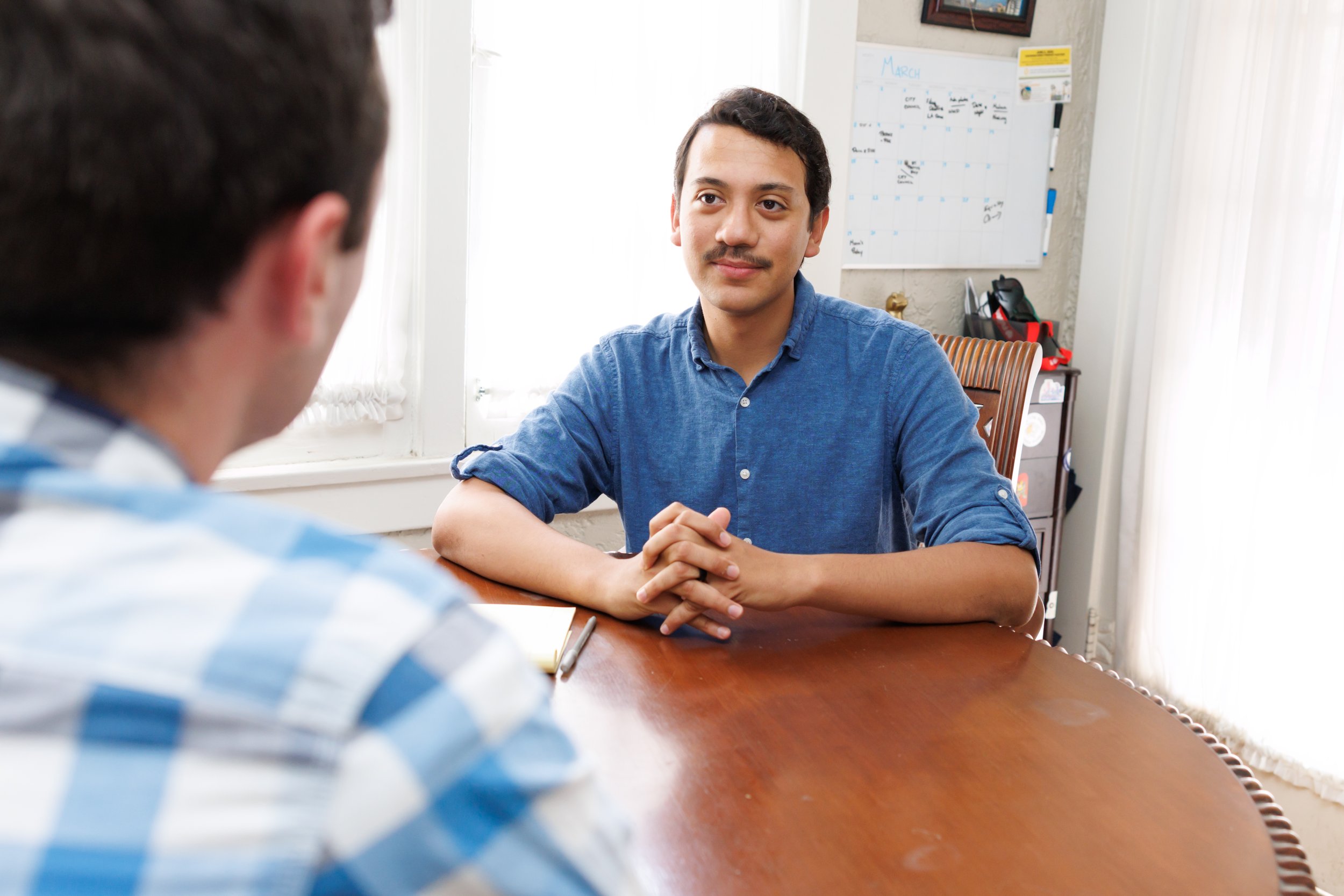 A man with dark hair and a mustache wearing a blue shirt in a meeting, with a calendar on the wall behind him and a desk with office supplies to his right. An out-of-focus person is in the foreground.