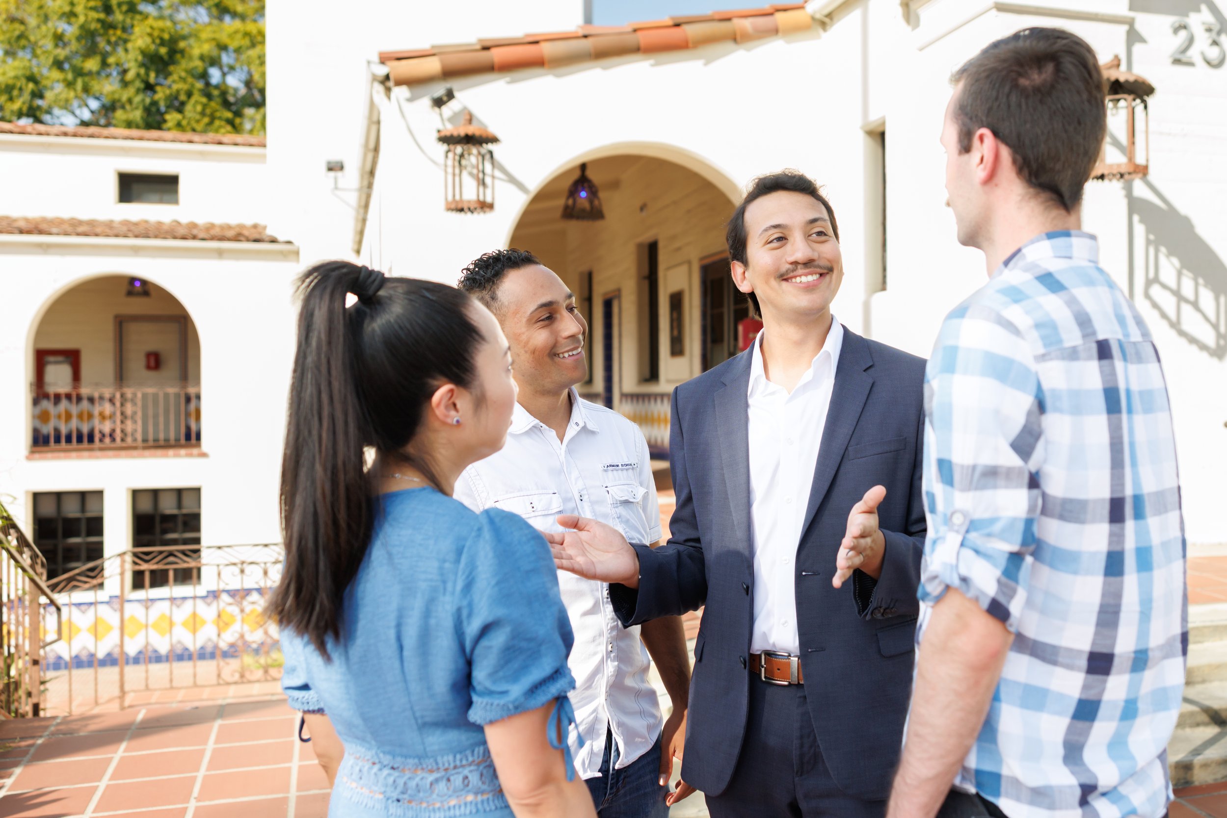 A group of four young adults, two men and two women, are talking and shaking hands outside a white building with Spanish-style tiled roof and arches, during daytime.