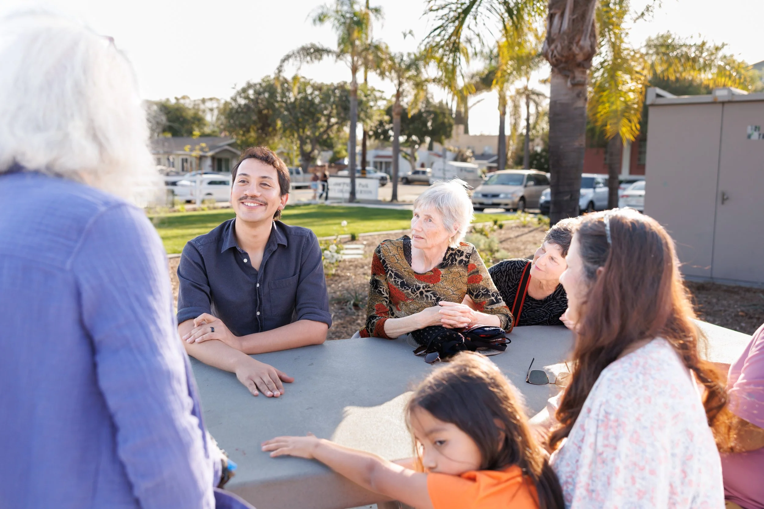 People gathered around a table outdoors, engaging in conversation during daytime in a park with trees and parked cars in the background.
