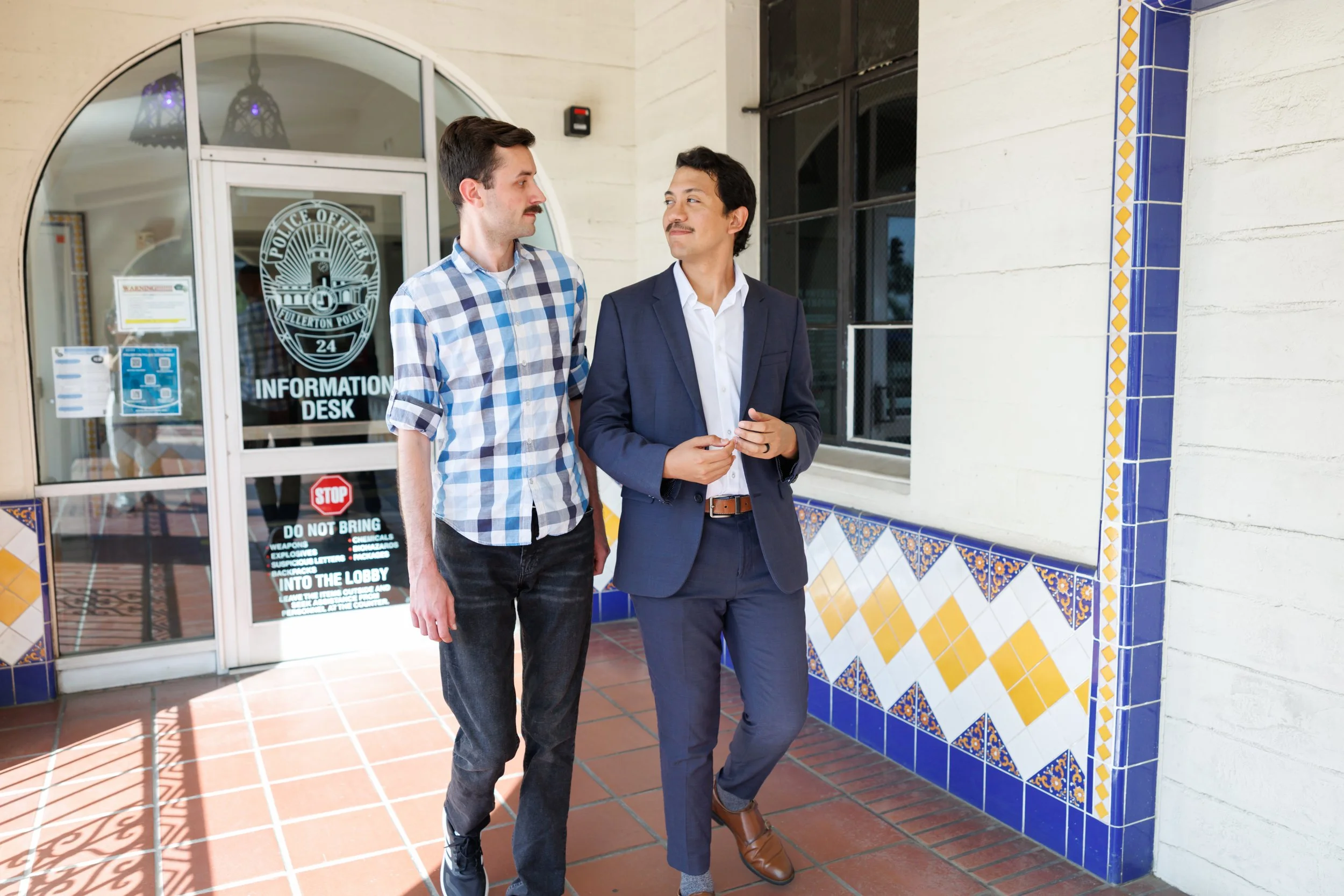 Two men standing outside a police station, engaging in conversation. The man on the left has light skin, dark hair, a mustache, and is wearing a blue and white checkered shirt with rolled-up sleeves. The man on the right has medium skin, dark hair, a