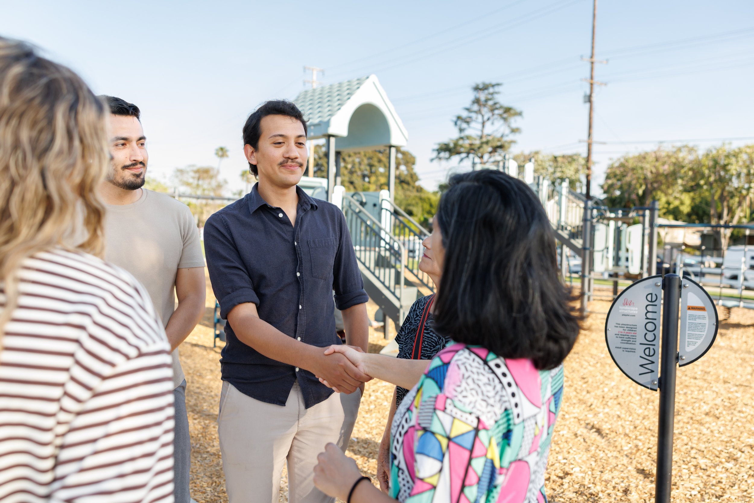 A group of five people, including a young man in a navy shirt shaking hands with a woman in a colorful dress, at a playground or park during daytime with trees and power lines in the background.