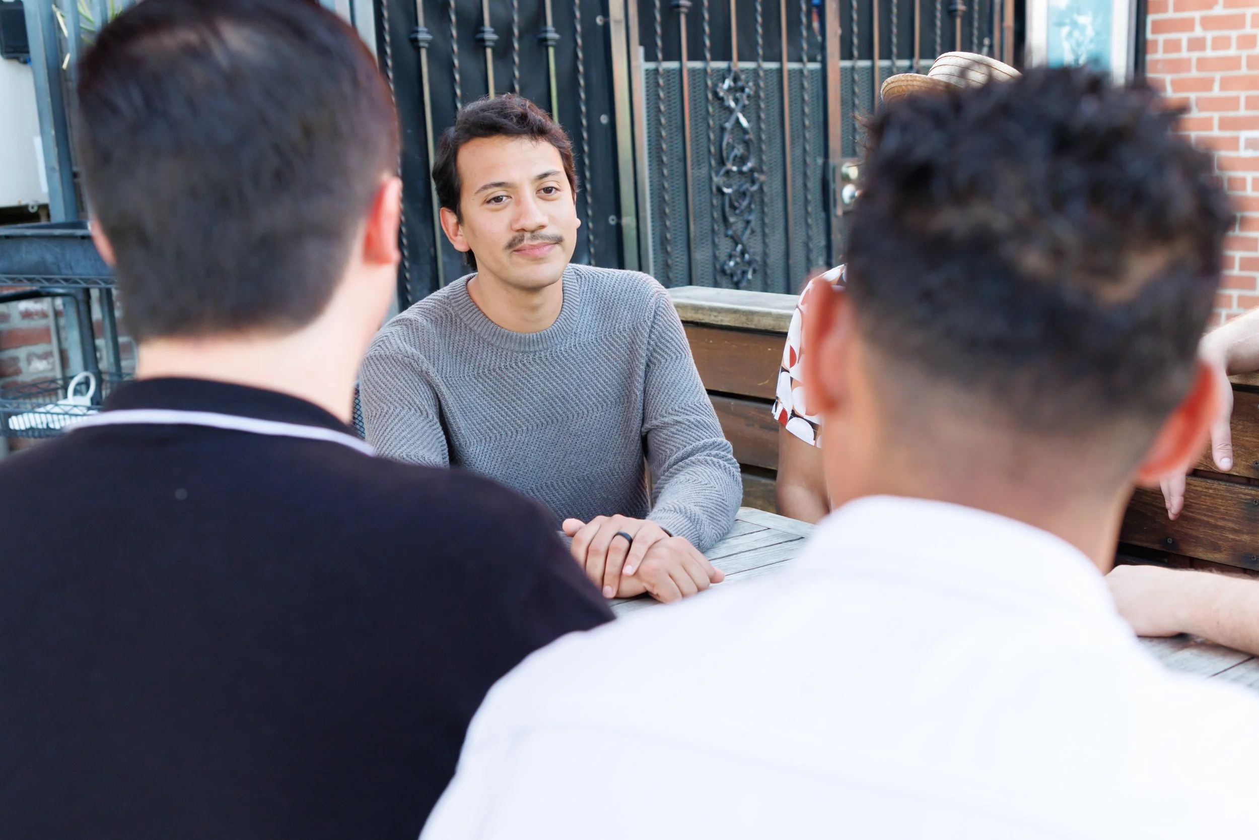 A man with dark hair and a mustache sitting at a table outside, facing three other people whose backs are visible, with a black metal fence and brick wall in the background.
