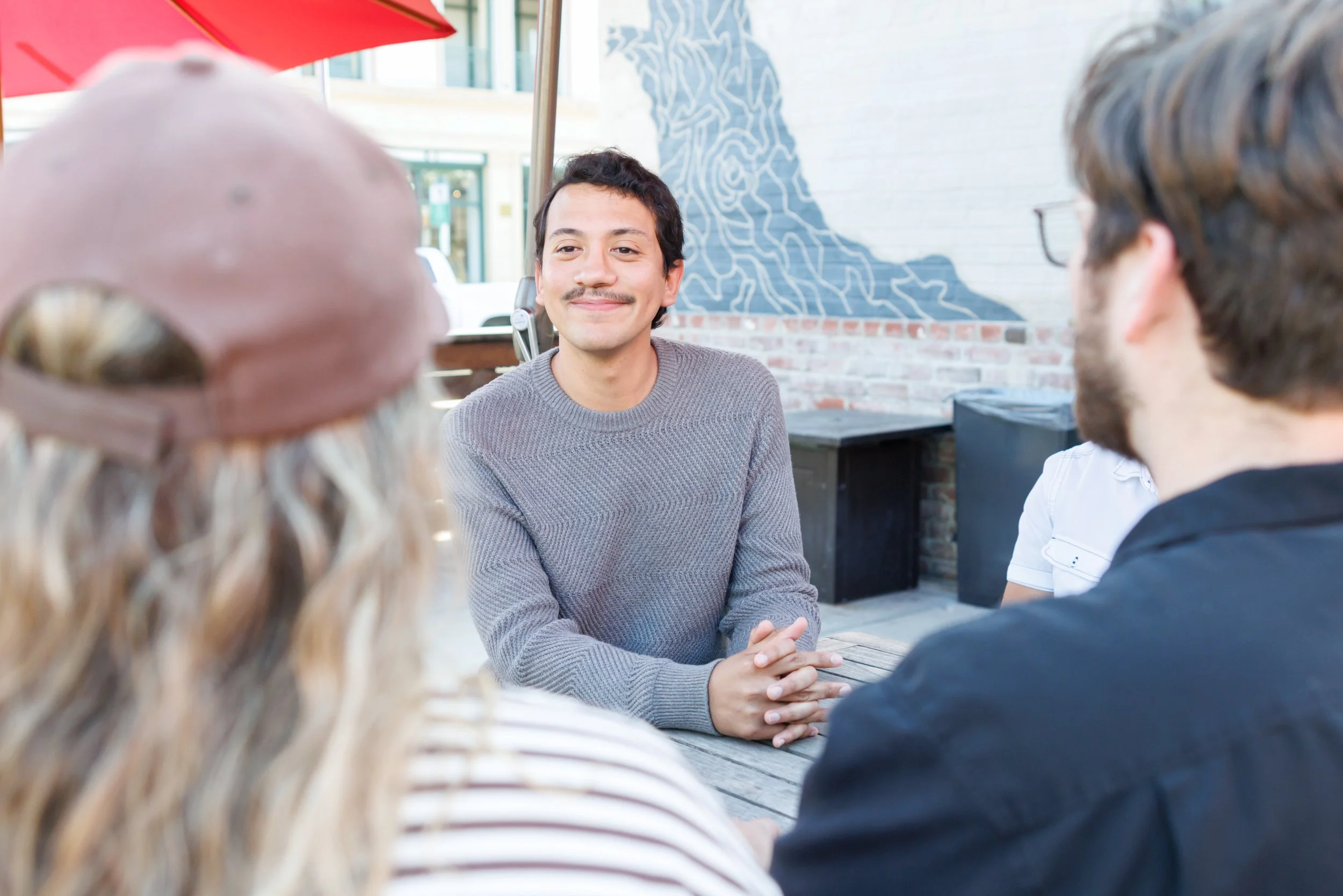 A young man with dark hair and a mustache smiling while engaging in conversation with a group of friends at an outdoor cafe.