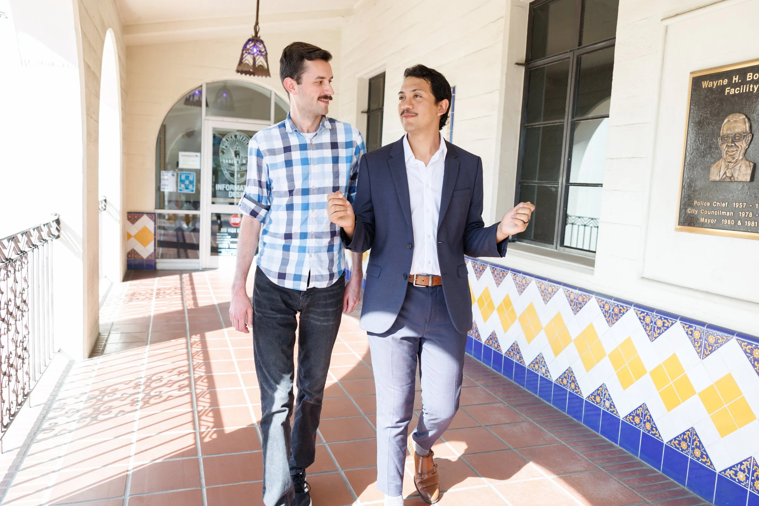 Two men walking and talking in a bright hallway with decorative tiles and a plaque on the wall.