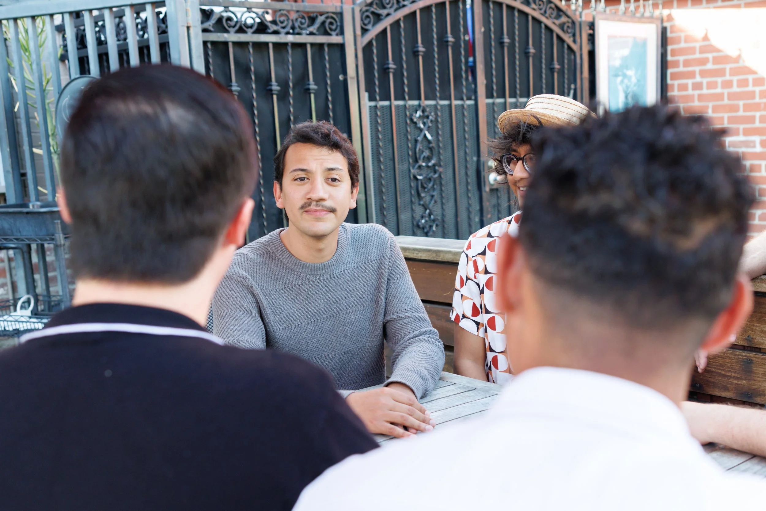 Group of five young adults sitting around an outdoor table, engaged in conversation. Four are visible from the back and side, one facing forward with light skin, dark hair, and a gray sweater.