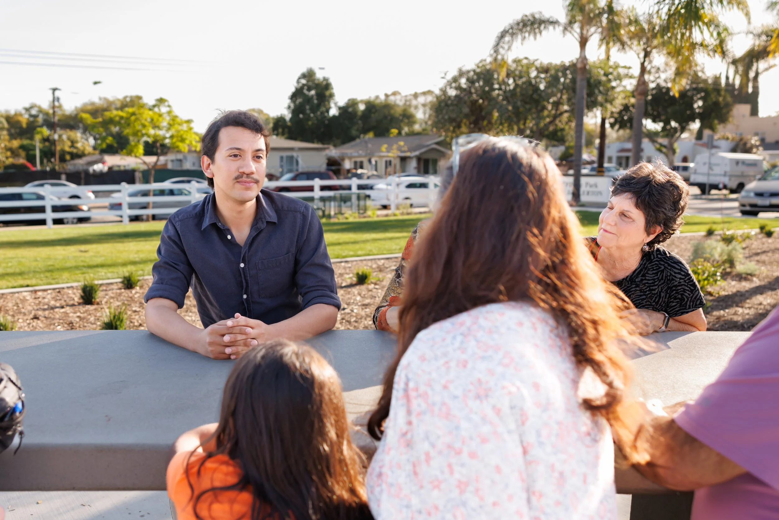 A young man with dark hair and a mustache speaking to a group of women sitting at a table outdoors in a park with trees and houses in the background.