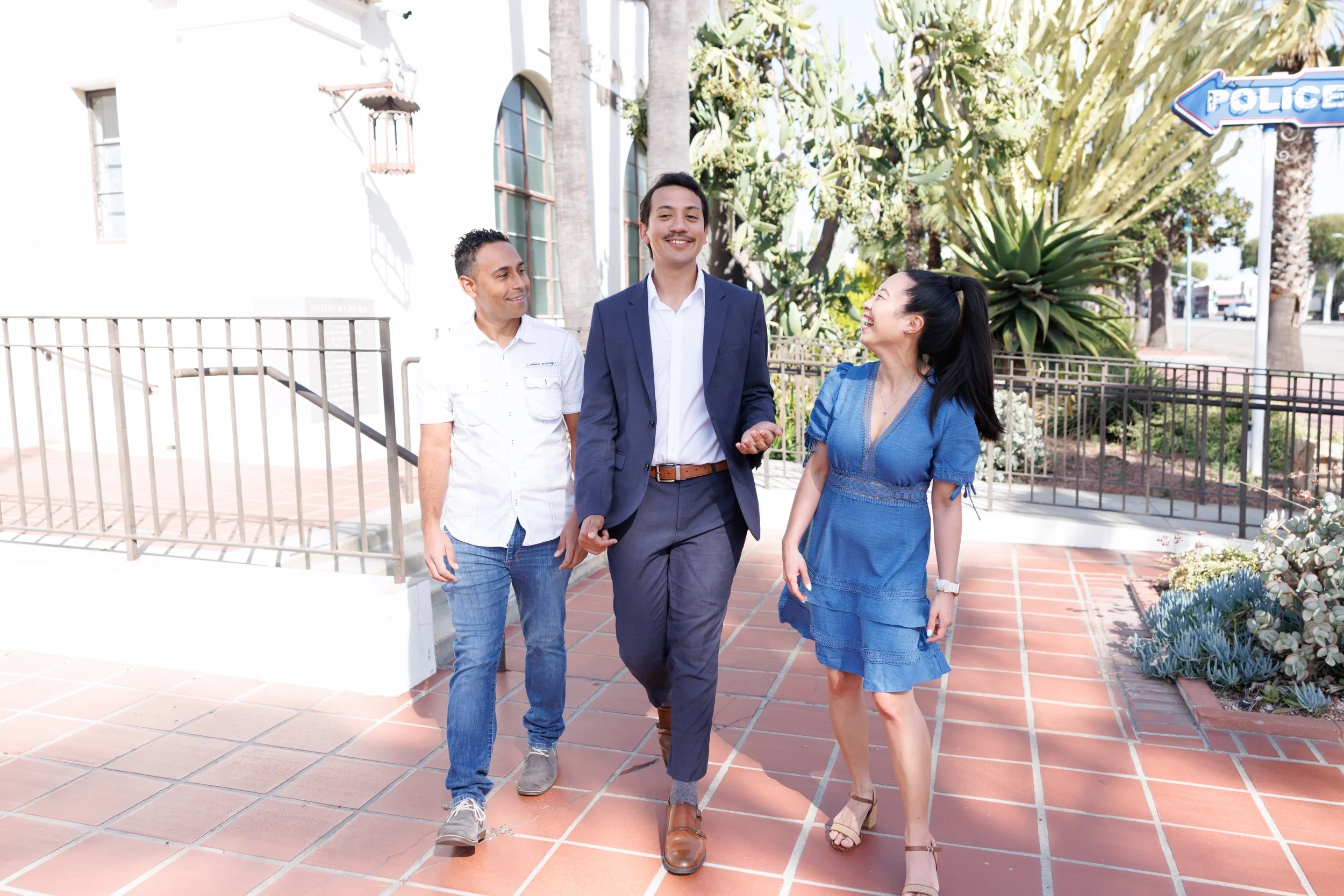 Three people, two men and one woman, walking and talking outdoors near a building with cactus and plants, with street and police sign in the background.
