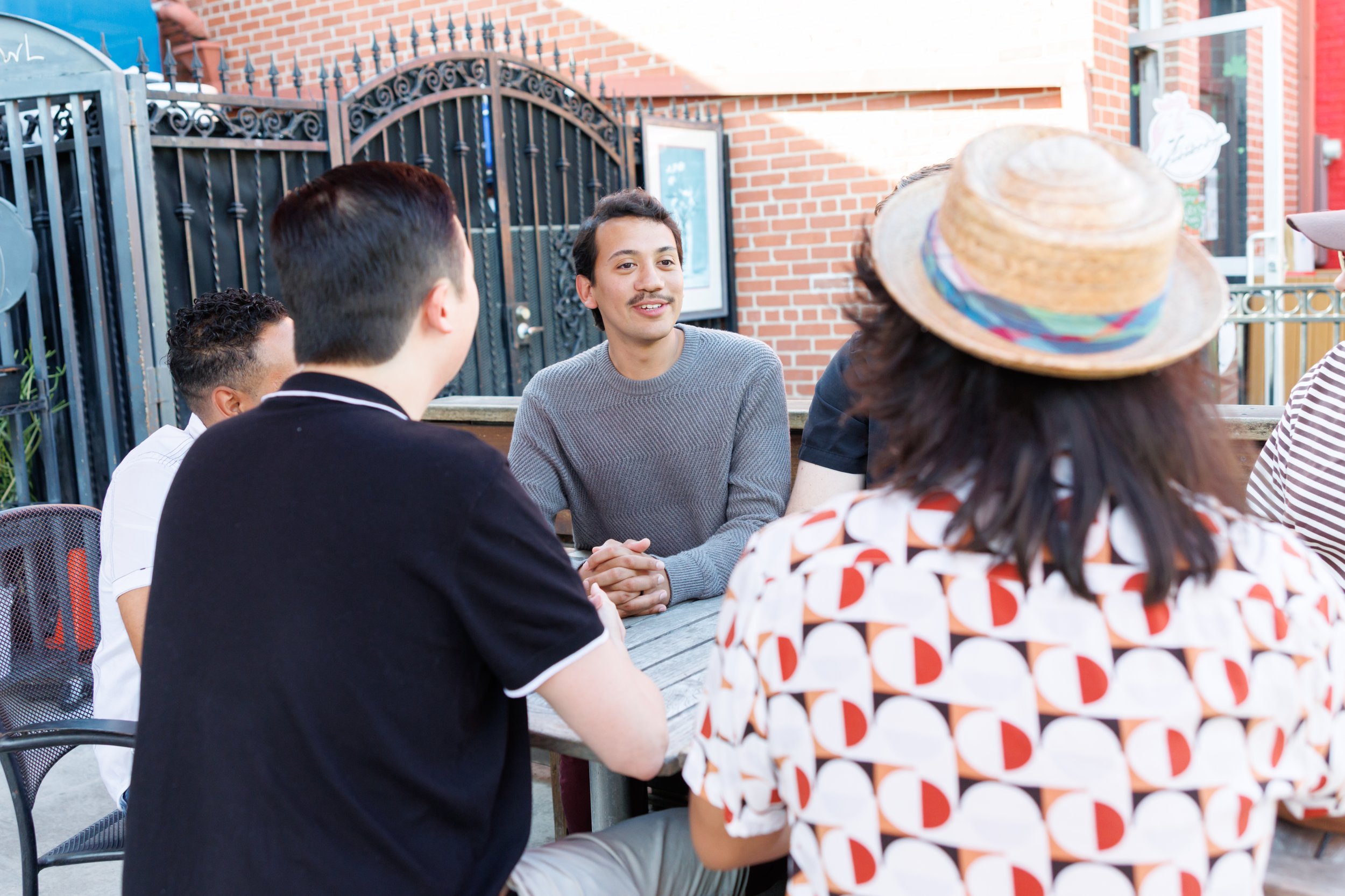 Group of people sitting outdoors at a table, engaged in conversation. A man with dark hair and a mustache is in focus, smiling while talking to others. Some people are wearing patterned clothing and a woman is wearing a straw hat.