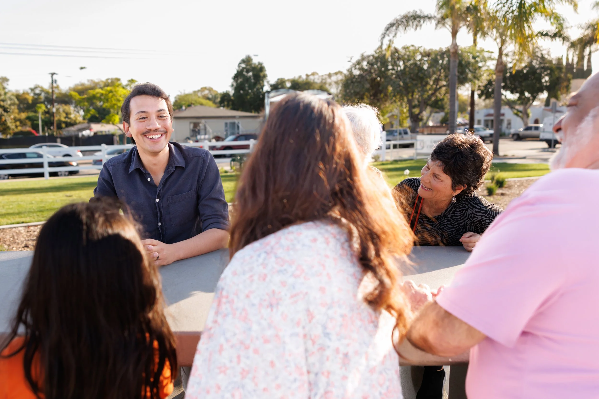 A group of people sitting outdoors on a sunny day, smiling and engaging in conversation. A man with a mustache in a dark shirt is prominently smiling. Others, including a woman with dark hair and a man with gray hair and a pink shirt, are part of the