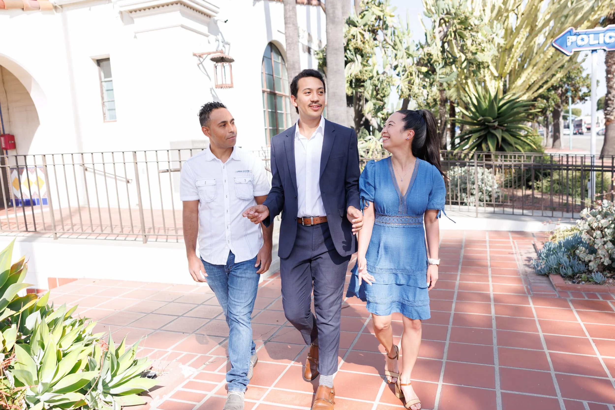 Three people walking outdoors in a sunny setting, with two men and one woman smiling and engaging in conversation. The man in the center is dressed in a dark blue suit, while the other man is wearing a white shirt and jeans, and the woman is in a blu