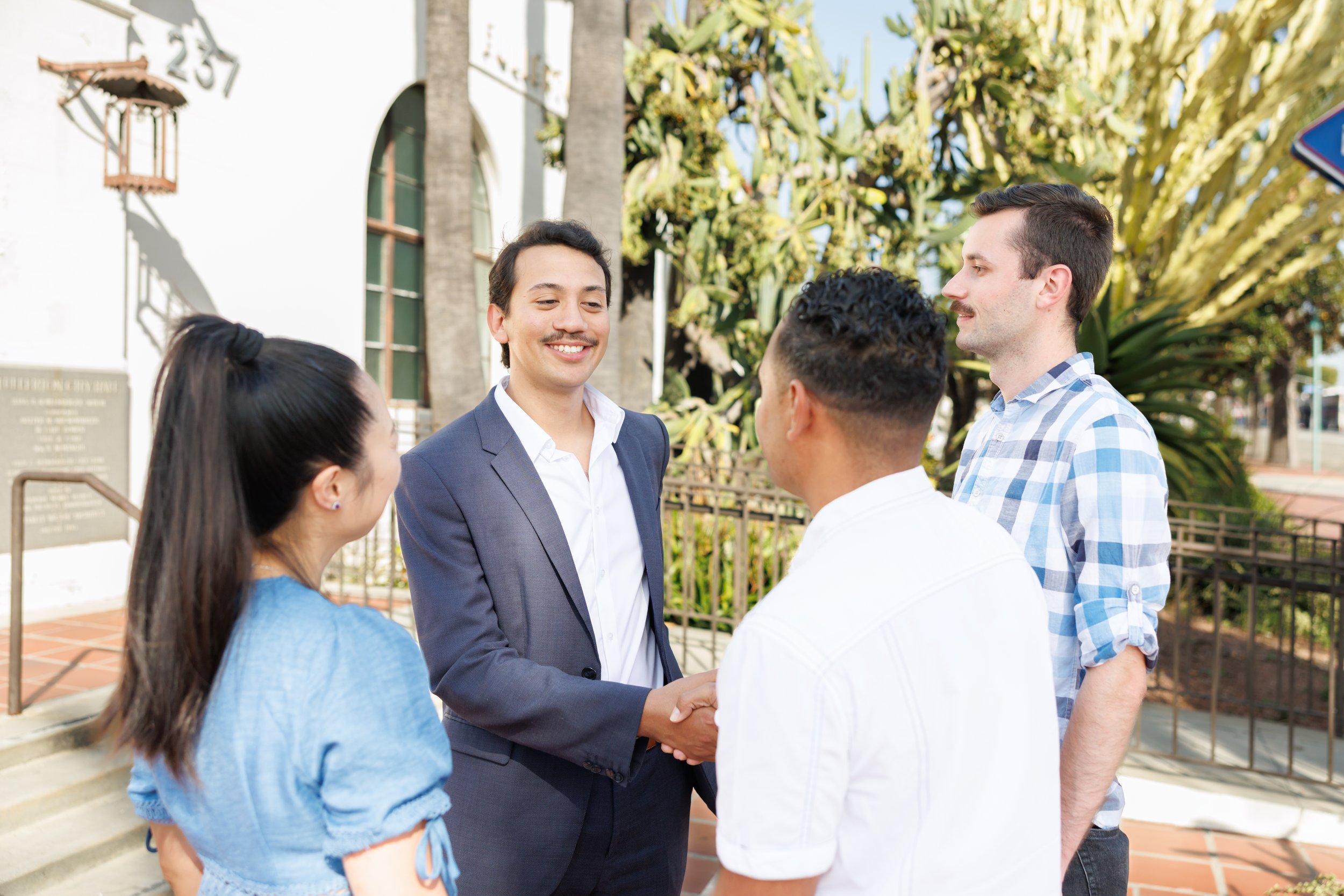A group of five diverse young adults gathered outdoors, engaging in a handshake and conversation with a man in a navy suit. The scene is bright with trees and a white building in the background.