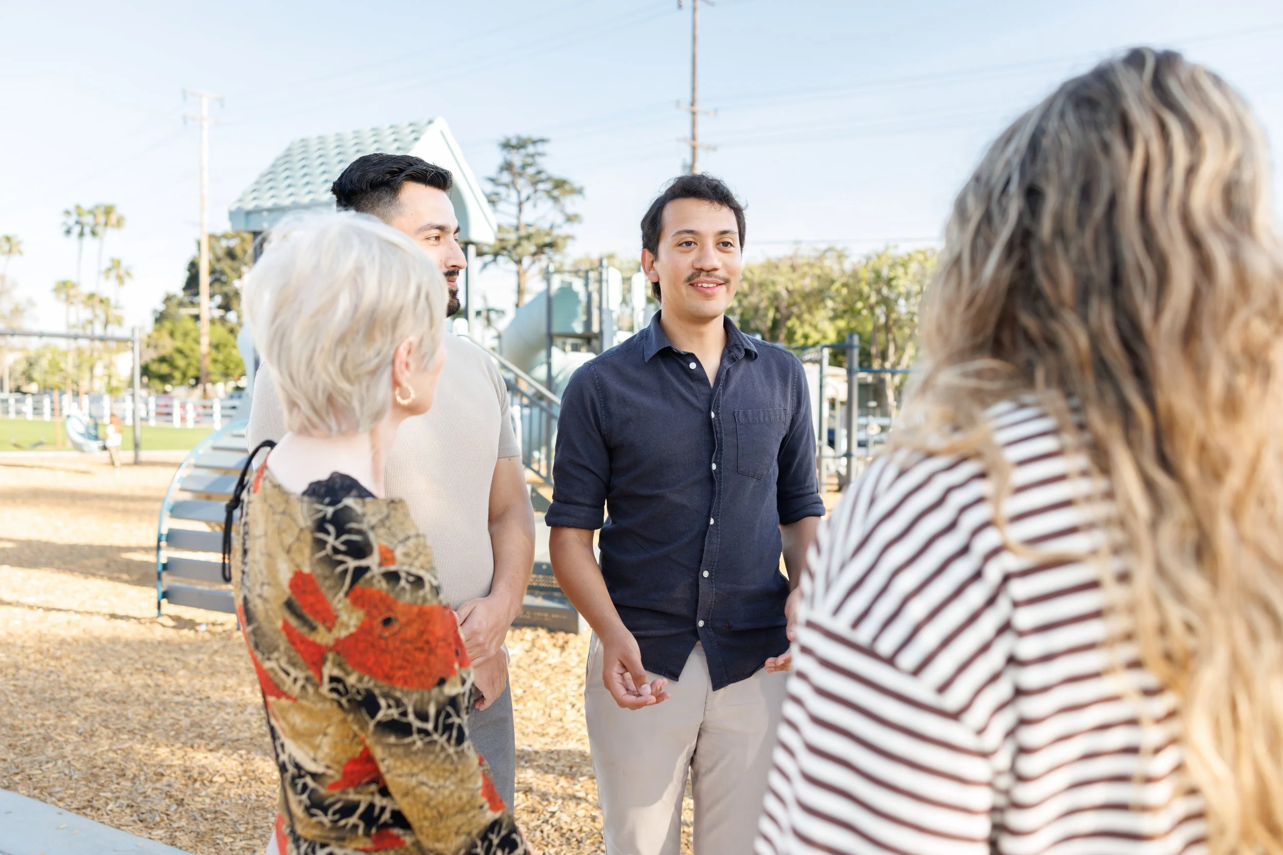 A group of four people standing outdoors, engaged in conversation on a sunny day. The background features trees, utility poles, and a park or playground area.
