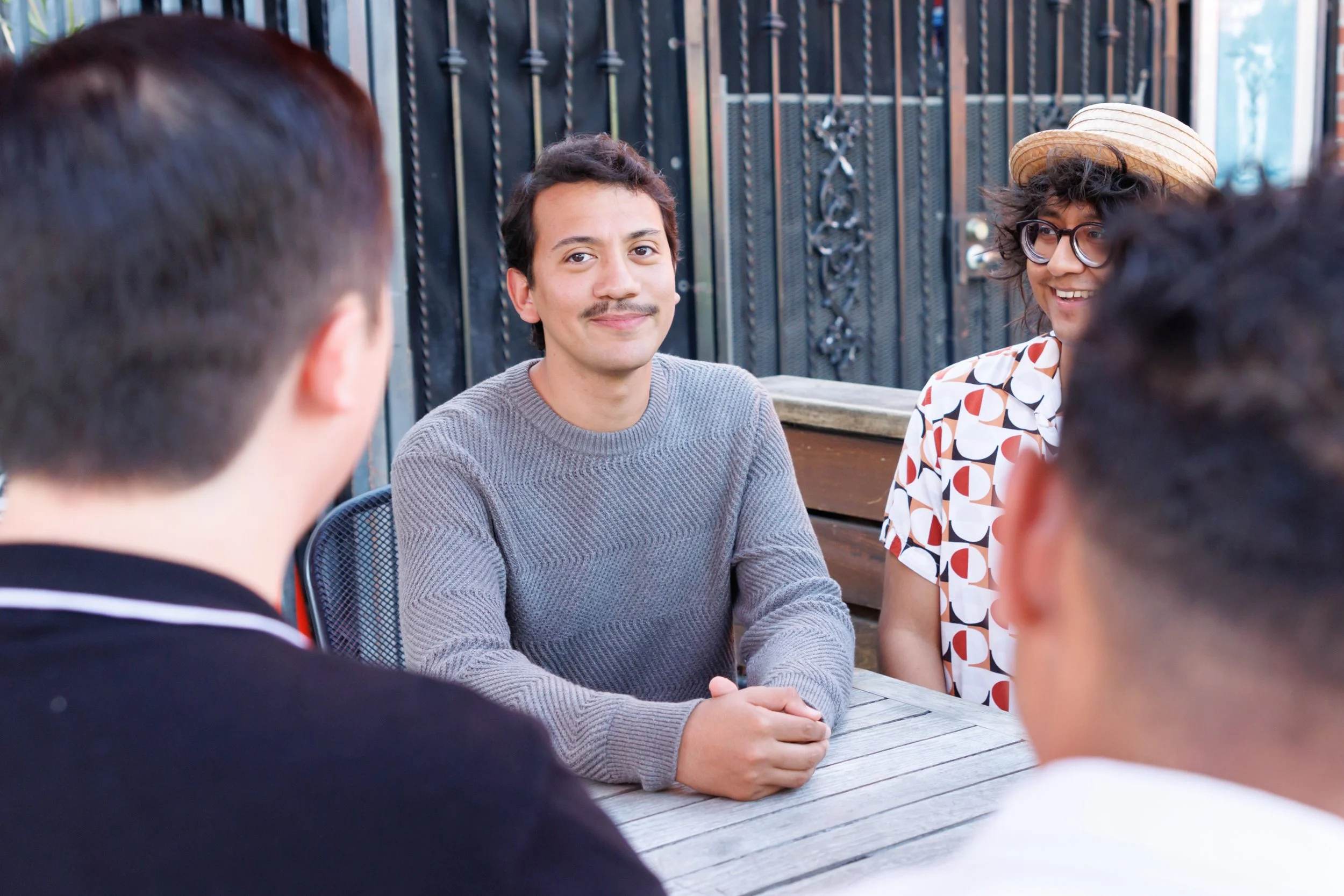 A group of young people sitting around a wooden table outdoors, engaged in conversation. One person in the middle is smiling, wearing a gray sweater, with others listening. The background features a black wrought iron fence.