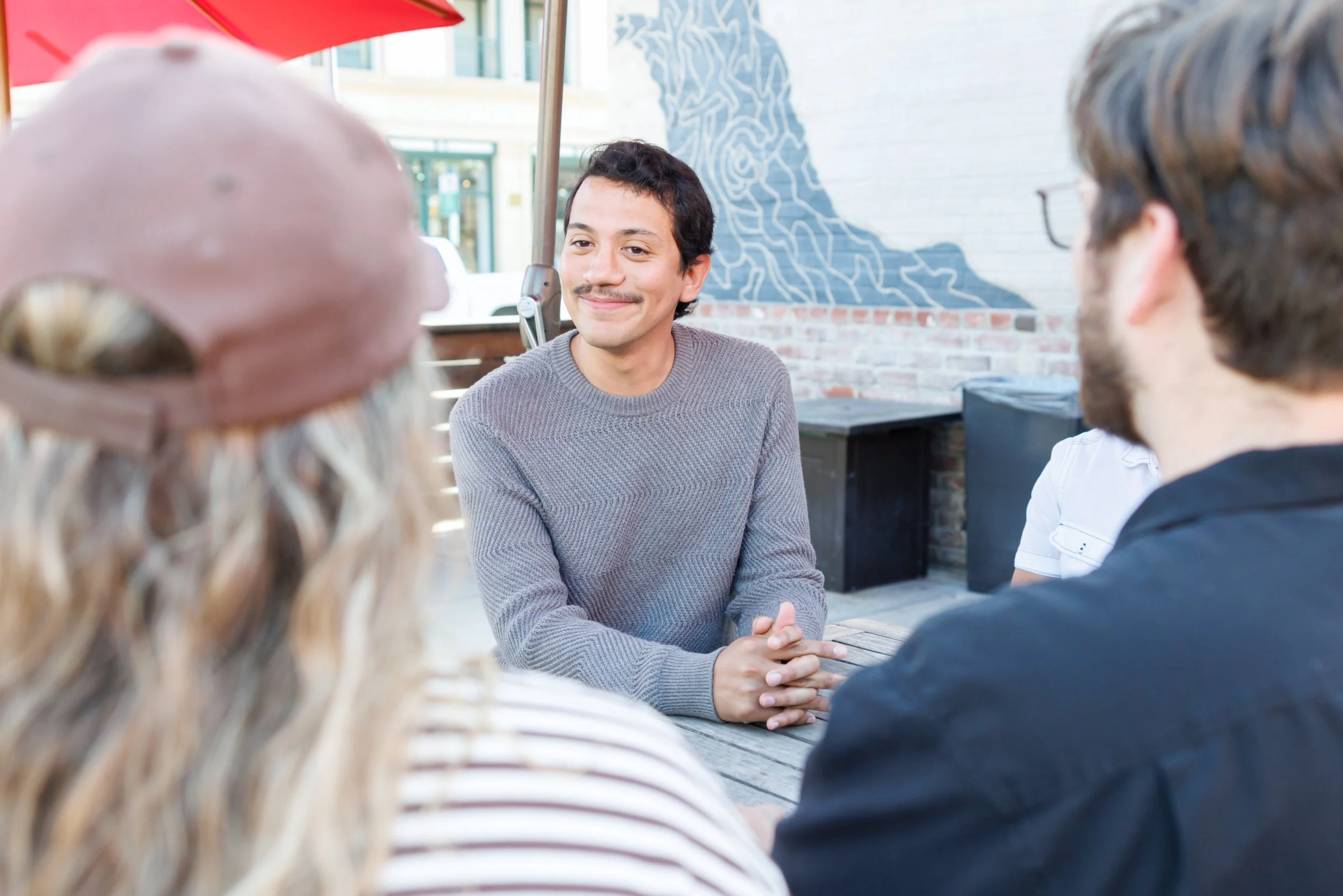 A man with dark hair and a mustache smiling while sitting with a group of people outdoors at a restaurant or cafe, with an abstract mural on a wall behind him.