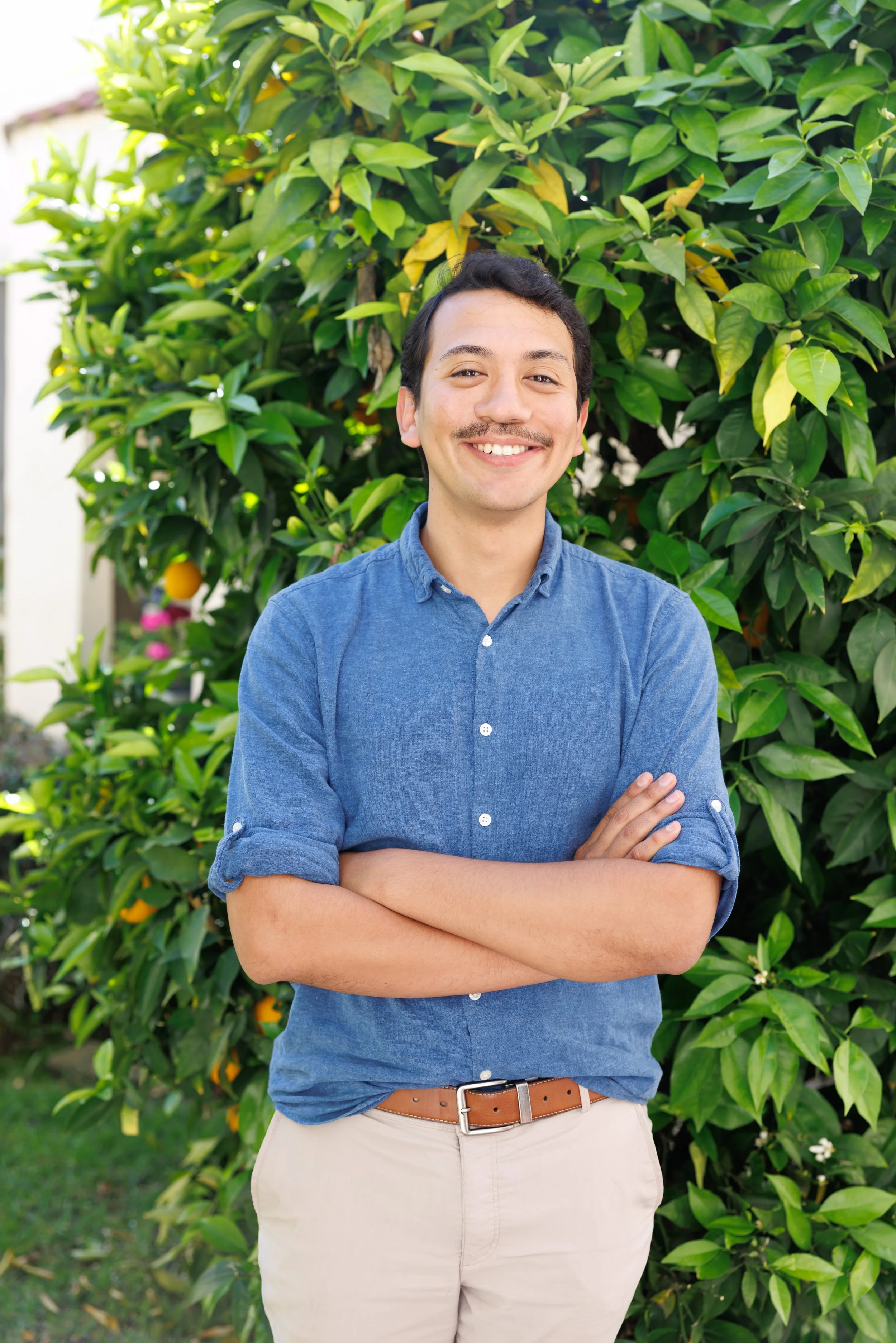 A young man with dark hair and a mustache smiling and standing outdoors with arms crossed in front of a tall green shrub with yellow fruit.