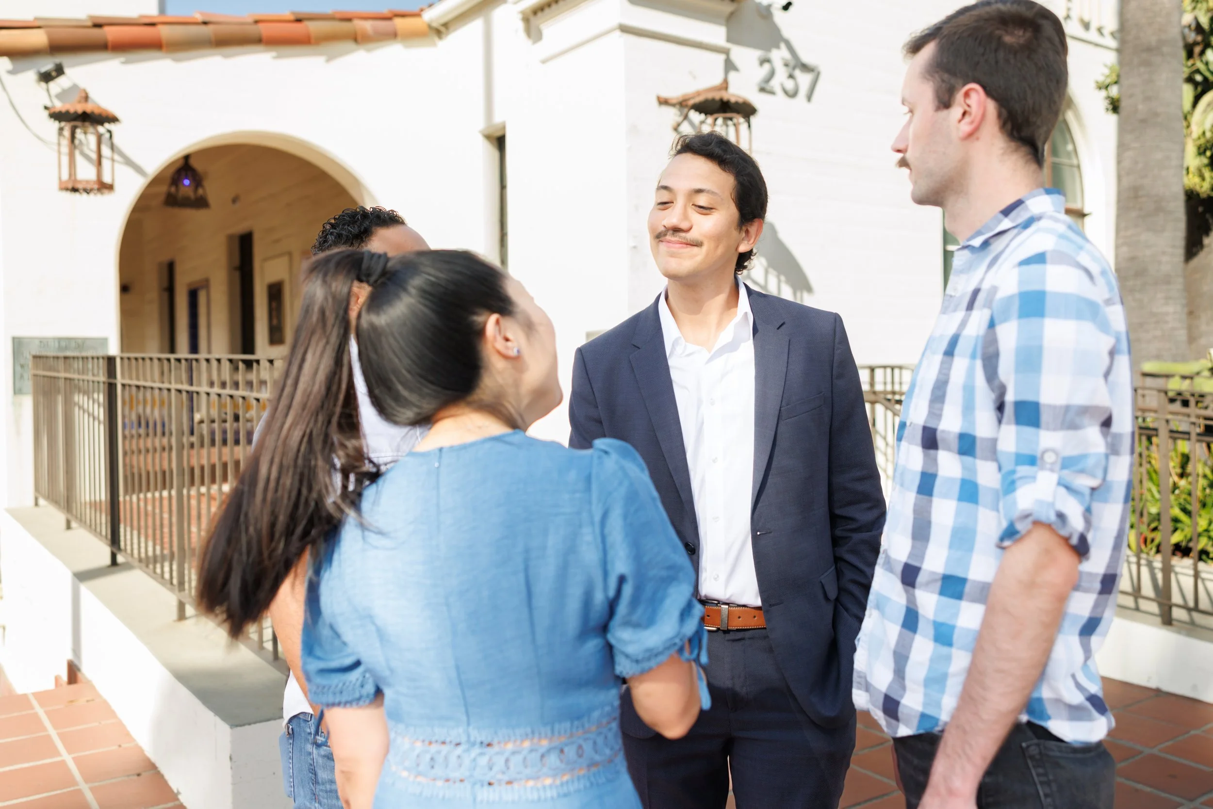 Group of five people engaged in conversation outdoors, standing in front of a white building with the number 237 on it. The group includes two men and three women, with one woman wearing a blue dress, another woman with a ponytail, and a man in a blu