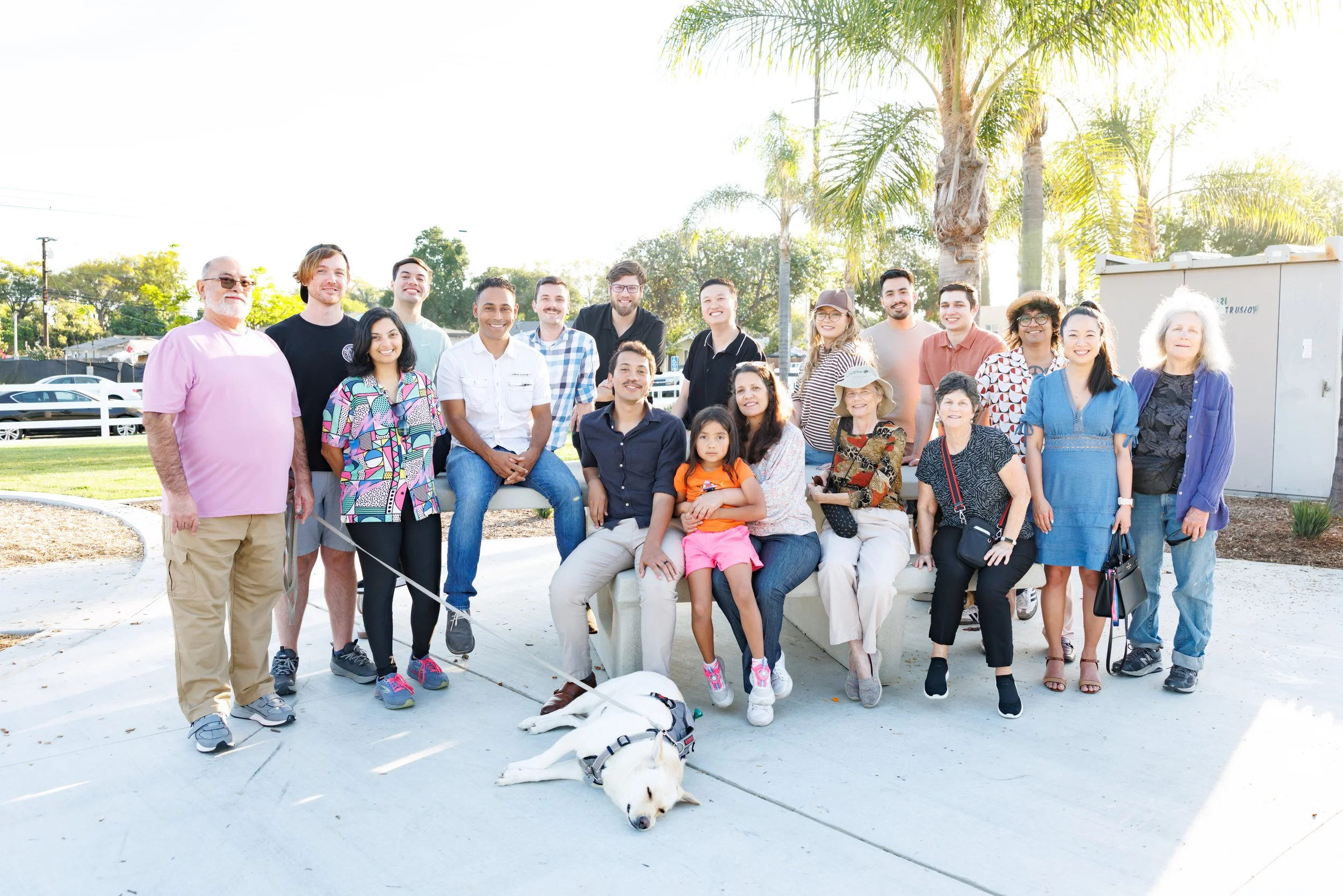 A diverse group of 18 people, including children and seniors, standing outdoors on a sunny day with palm trees in the background. They are smiling and posing for a group photo, with a white dog lying on the ground in front of them.