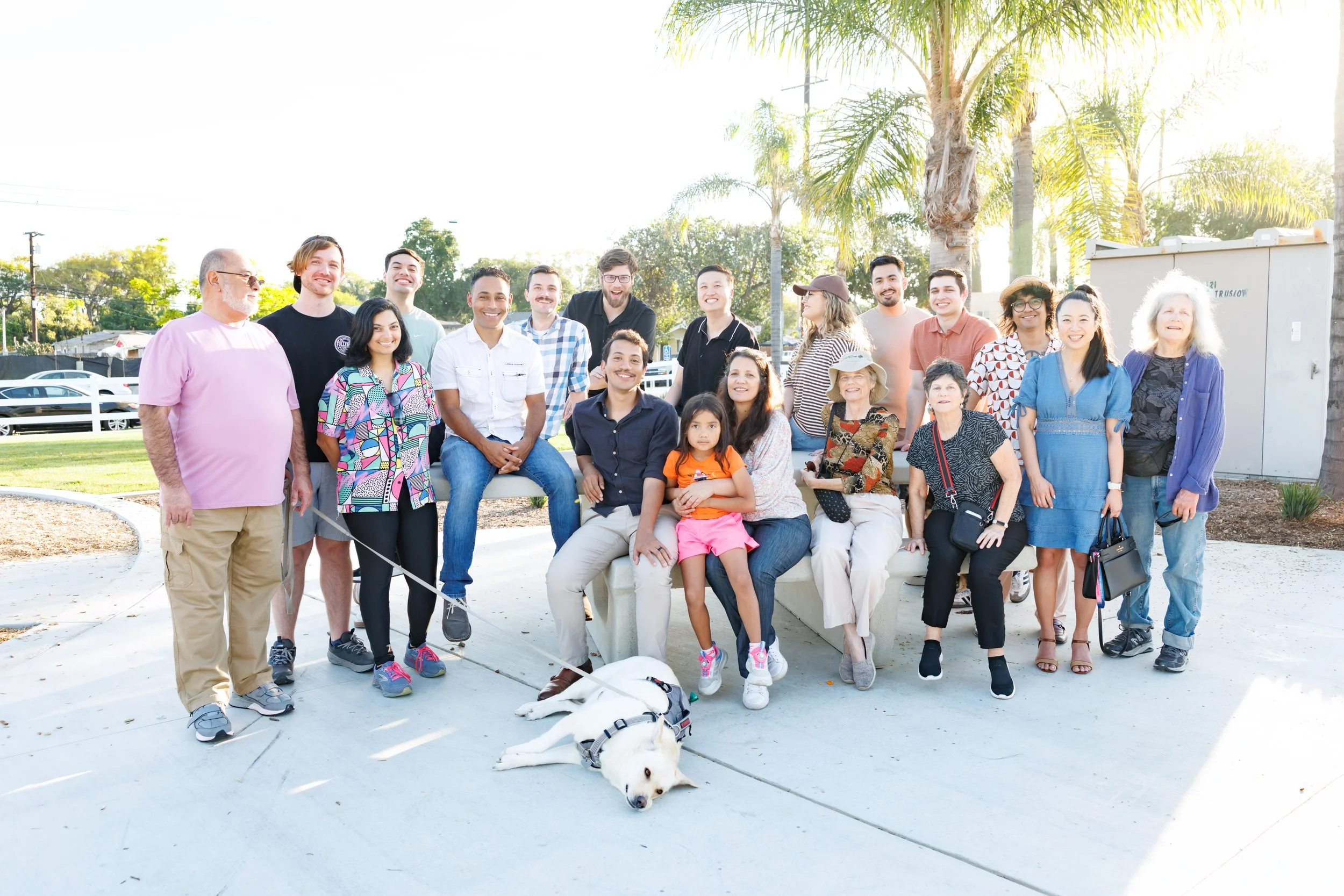 Group of diverse people smiling and posing outdoors in a park with palm trees and green foliage, with a white dog lying on the ground in front.