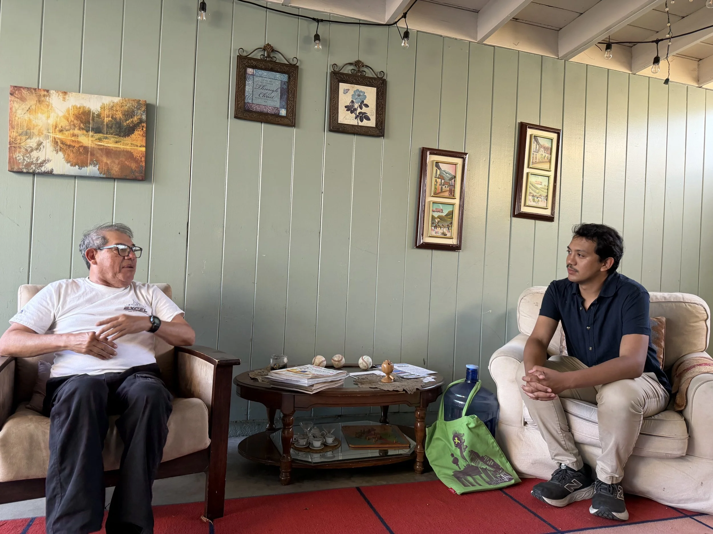 Two men are sitting on separate sofas in a cozy room with green paneled wall decorated with framed artwork. They appear to be engaged in conversation, with a coffee table between them holding books, magazines, and a small decorative item. The room has a ceiling with exposed beams and hanging light bulbs, and a red rug sits on the floor. One man is older with glasses and grey hair, wearing a white t-shirt, while the other is younger with dark hair, wearing a dark shirt.