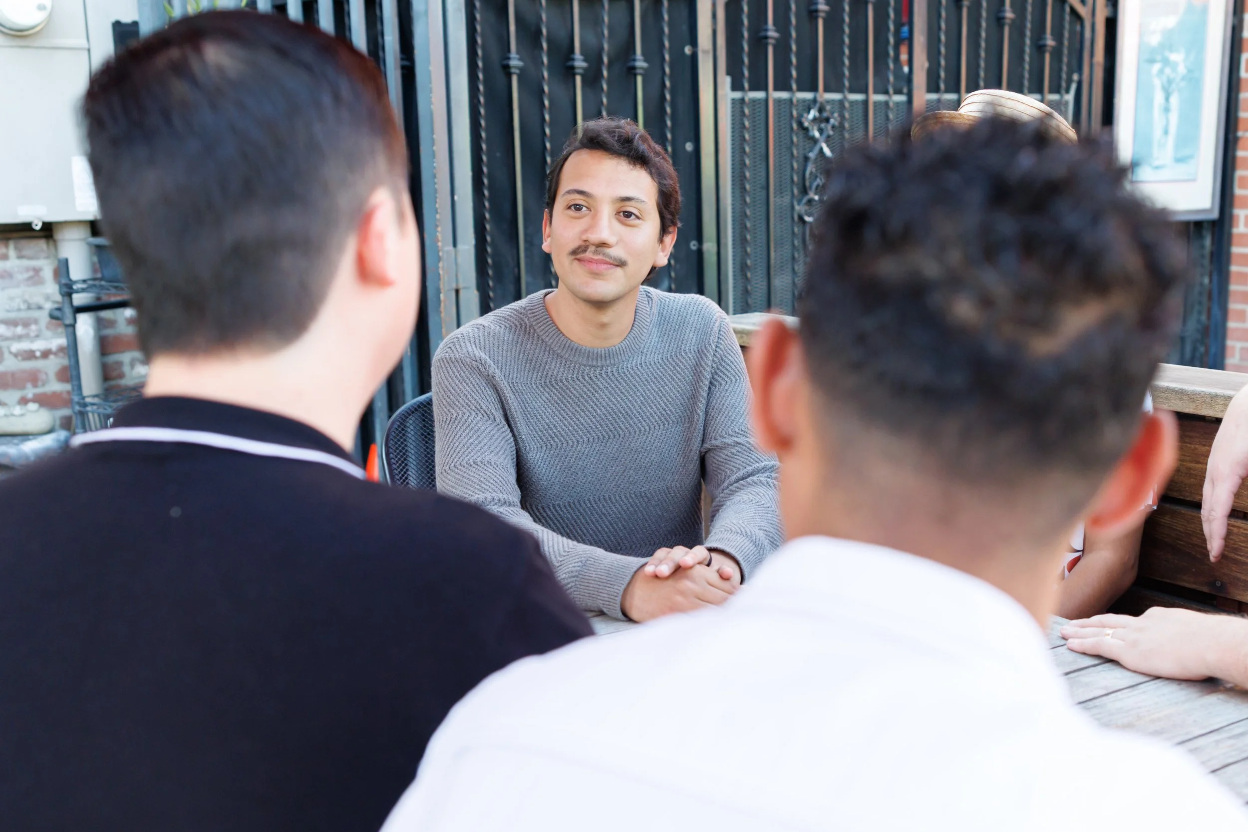 A young man with short dark hair and a mustache, wearing a gray sweater, is sitting at a table outside, engaging in conversation with two people whose backs are visible. They are seated at a wooden table with a black metal fence and some hats hanging