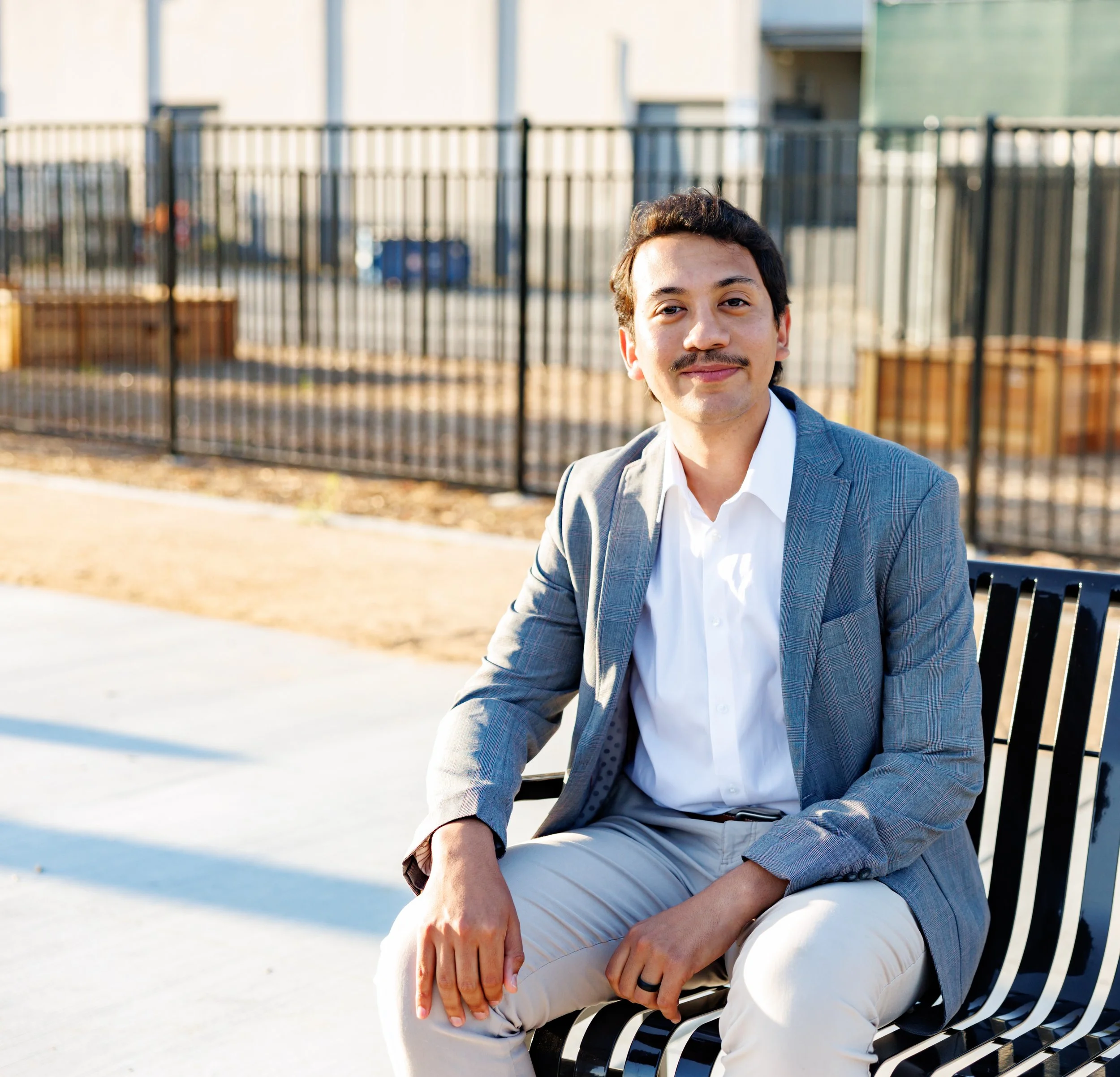 A young man with dark hair, wearing a light gray blazer, white shirt, and light-colored pants, sitting on a black metal bench outdoors near a fenced area. He's smiling, holding his left leg with both hands, with the sunlight casting warm light on his